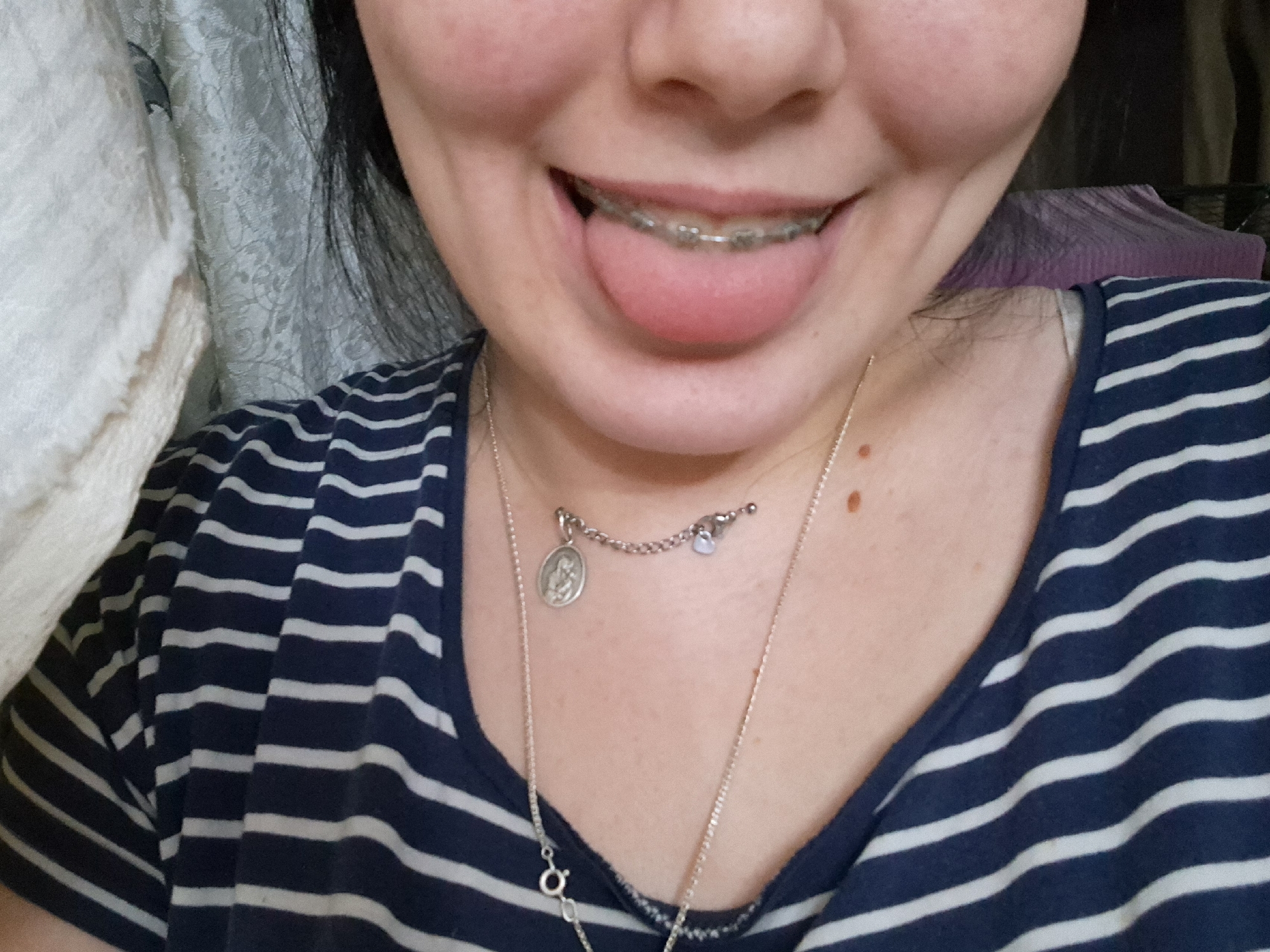 The image is a close-up, portrait-style photograph of a person's upper chest, neck, and mouth. The subject has dark hair that frames their face and is wearing a navy blue and white striped shirt. Two silver necklaces are visible; one is shorter with a circular pendant featuring an image and a small pale blue charm, and the other is longer and thinner. The subject's mouth is slightly open, with their tongue extended, revealing orthodontic braces on their teeth. The skin on the neck has a few small moles. The background is out of focus, showing what appears to be a textured fabric, possibly a curtain or bedspread, in light and darker colors. The lighting suggests an indoor setting, and the overall mood is casual and playful due to the tongue gesture and visible braces. There is no visible text in the image.