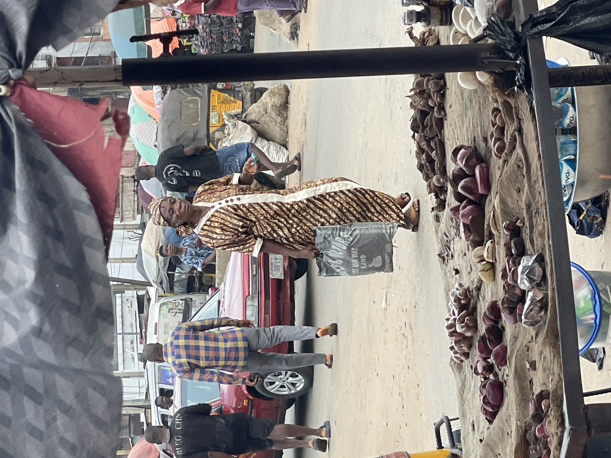 This image depicts a bustling street market in Lagos, Lagos State, Nigeria. In the foreground, a market stall covered with a burlap-like fabric displays various goods including piles of reddish and yellowish kola nuts, dried dark brown roots or herbs, a cluster of white eggs, and small bottles. Underneath the stall, a silver bowl holds multiple "eva" branded bottled water, positioned next to a blue bucket.
Several people are in motion. In the midground, a woman wearing a brown, gold, and white patterned dress and a matching headwrap walks from right to left, carrying a dark grey shopping bag in her left hand. The bag features white text, partially legible as "M & MOTORS" and "105 MN WAY KANO." Behind her, a man in a black t-shirt with "BLEG CHN" printed on the back and black shorts walks away from the camera. Adjacent to him, another man in a blue, yellow, and red plaid long-sleeved shirt and grey trousers walks away, holding a phone in his right hand.
A red Toyota sedan with a visible license plate "LAGOS C306-ENO" is parked or slowly moving in the midground. Behind the sedan, a yellow auto-rickshaw (Keke Napep) branded "TVS" is partially visible. The background features more individuals and numerous open, colorful market umbrellas (blue, green, orange, red), indicating additional stalls. Older, multi-story buildings line the street. The ground is an unpaved or poorly maintained sandy-dirt surface. A dark grey patterned awning or tarp partially covers the top of the frame.