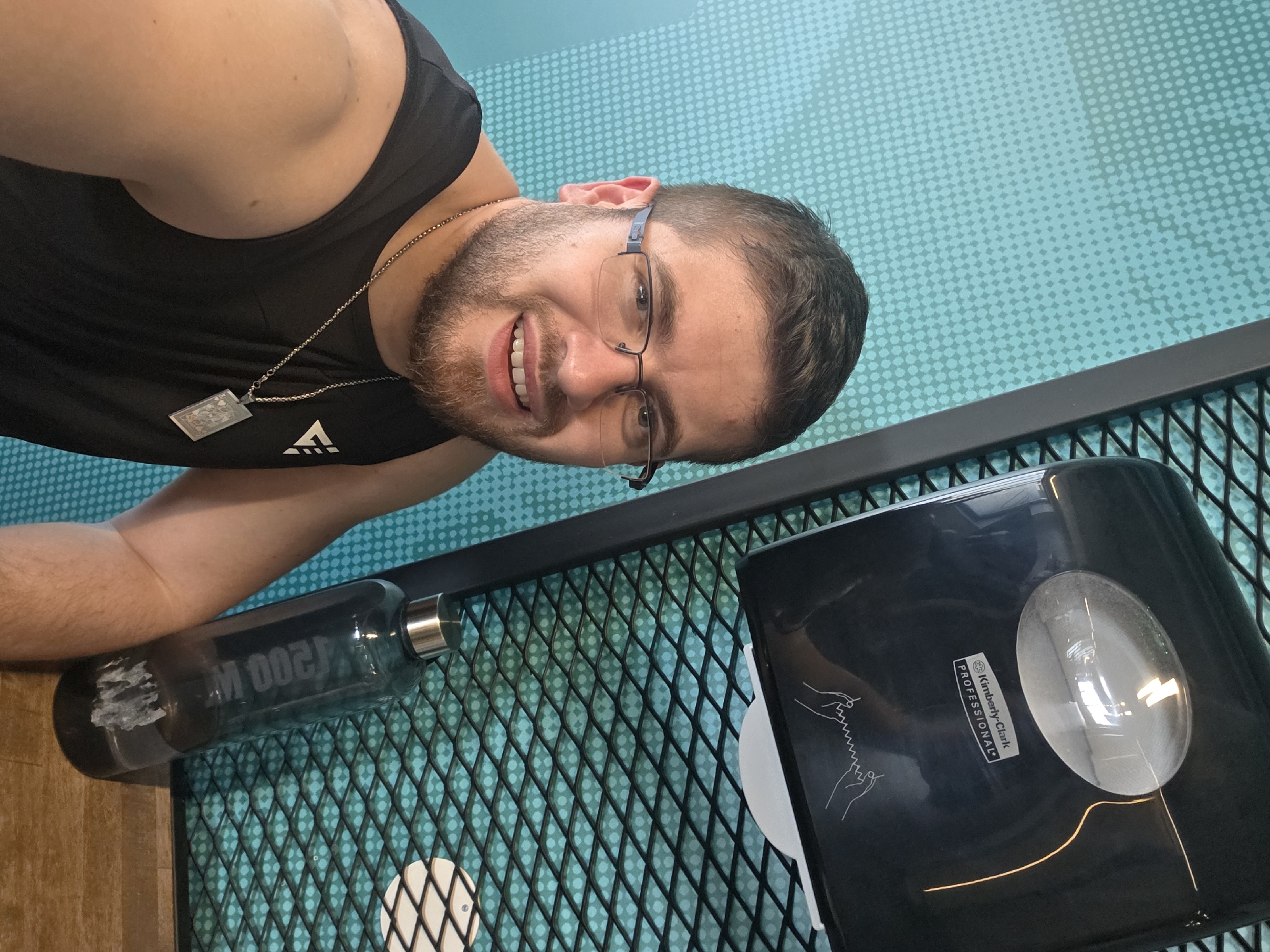 A selfie shows a man indoors, likely in a gym, in Plovdiv, Bulgaria. He wears glasses and a black tank top with a necklace. He is smiling at the camera. To his right are a water bottle and a Kimberly-Clark Professional paper towel dispenser resting on a black metal rack. The wall behind him is turquoise with a patterned design.