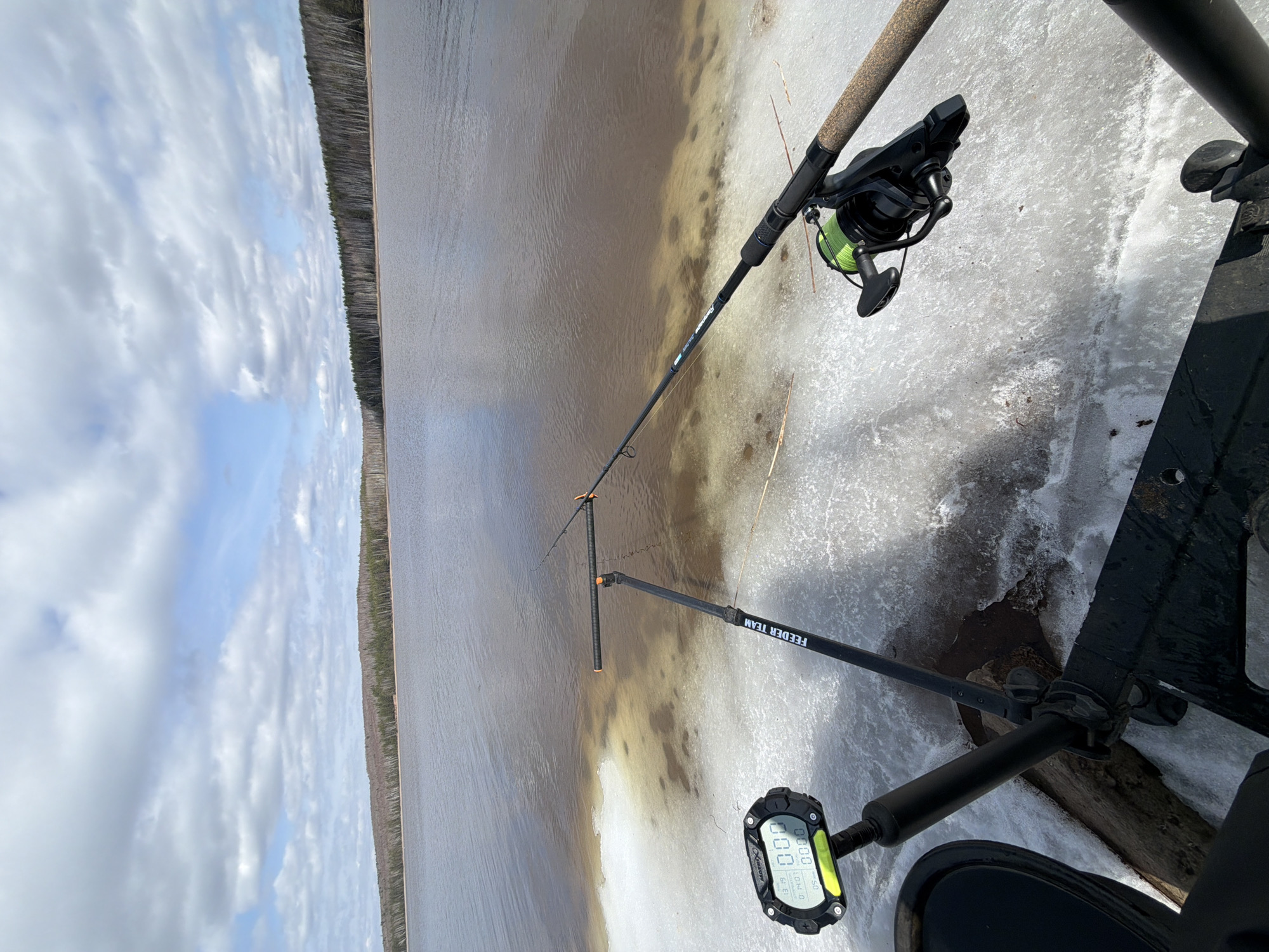 This image captures an outdoor fishing scene from an elevated perspective, likely on the bank of a lake or wide river in Chaykovsky, Russia, during a cold, transitional season like early spring or late autumn. The sky, filling the upper portion of the frame, is partly cloudy with large white clouds interspersed with patches of blue.
A distant, dense forest line stretches across the horizon, separating the sky from the body of water. The water itself shows a mixture of conditions; areas closest to the viewer are covered in melting ice and slush, while the central part of the water appears open and relatively calm, with a murky, brownish-grey color.
In the foreground, a black fishing rod, characteristic of a feeder rod, is set up and extends into the open water. It is equipped with a black spinning reel spooled with vibrant neon green fishing line. The rod rests on a black feeder rod rest that prominently displays the white text "FEEDER TEAM". Below and to the right of the rod, a digital device, possibly a fish counter, timer, or depth finder, is visible. The device's screen shows the brand name "X-Braid" and various numerical readings such as "0000", "SU", "0.01M", and "01H:07M", along with "01.3". Part of a dark fishing platform or chair base is also visible in the lower right corner, suggesting a comfortable setup for fishing. The scene evokes a sense of quiet patience and dedication to the sport amidst a rugged natural environment.