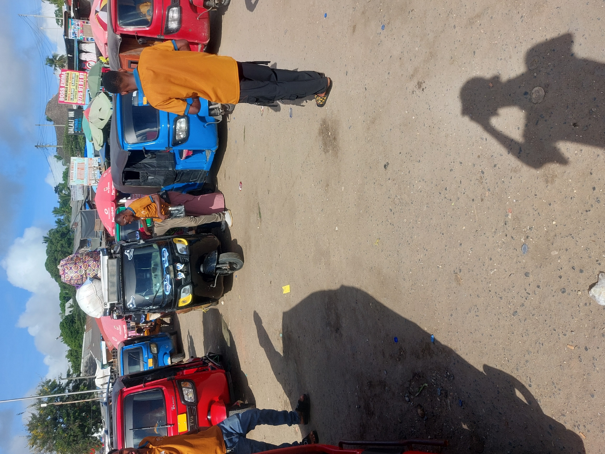 The image captures a dusty roadside scene in Tanzania. It is populated by a line of colorful three-wheeled vehicles, likely auto-rickshaws or "bajajis," in shades of red, blue, and black. People are seen standing near the vehicles, some wearing bright yellow shirts. The background includes buildings with signs written in Swahili, which suggest the location is urban, potentially the city of Dar es-Salaam. One of the signs advertises properties and contains contact information. The sky is partly cloudy, and the sun casts long shadows on the ground.