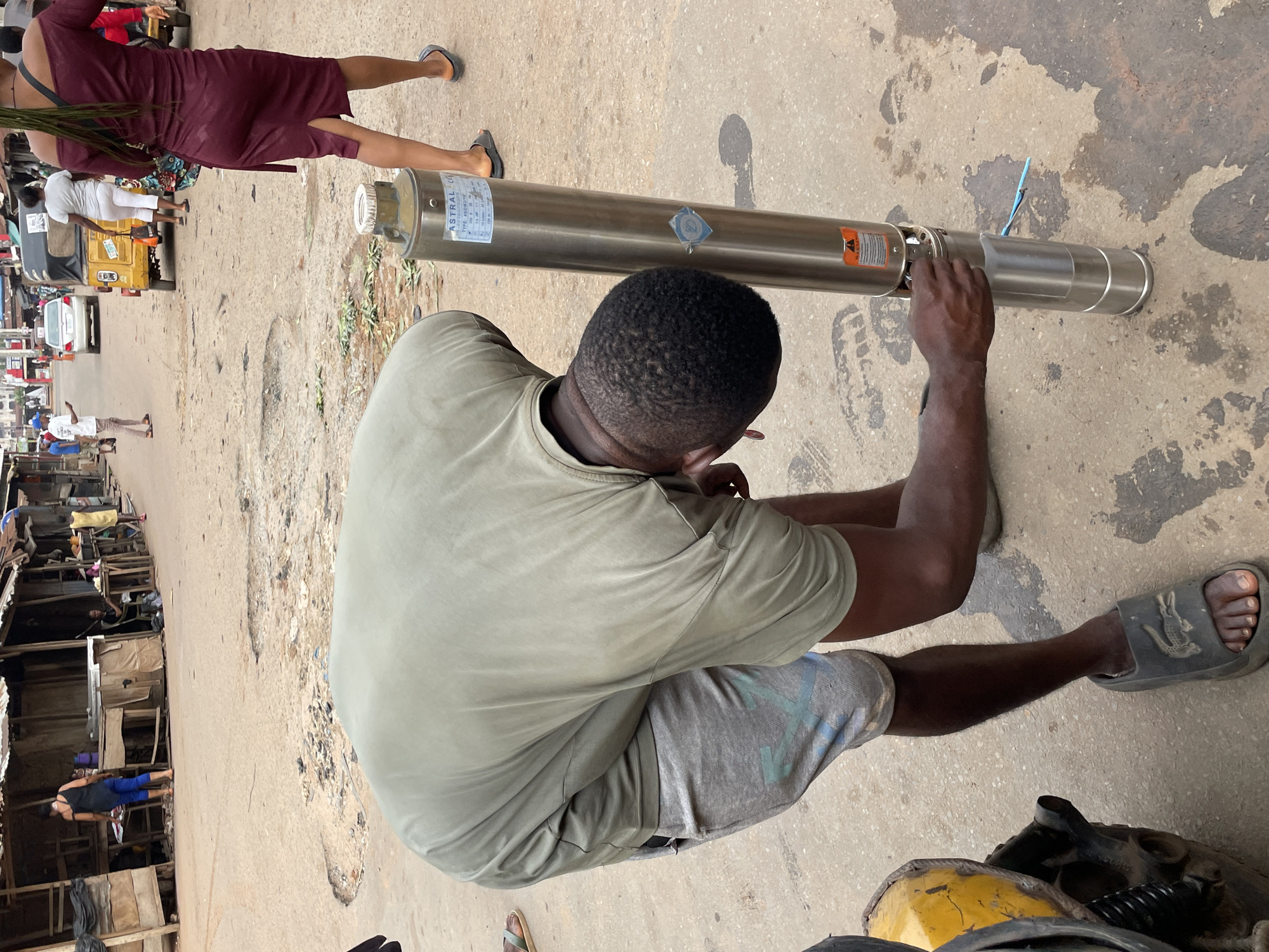 A man kneels on a concrete surface, manipulating the end of a long, cylindrical metal object labeled "ASTRAL." He wears a light green t-shirt, grey shorts, and dark grey sandals. To the left, a woman in a burgundy dress walks away from the camera. In the background, a bustling street scene includes a yellow taxi, various buildings, and pedestrians. The scene suggests a crowded, urban environment, indicative of a location in Onitsha, Nigeria.