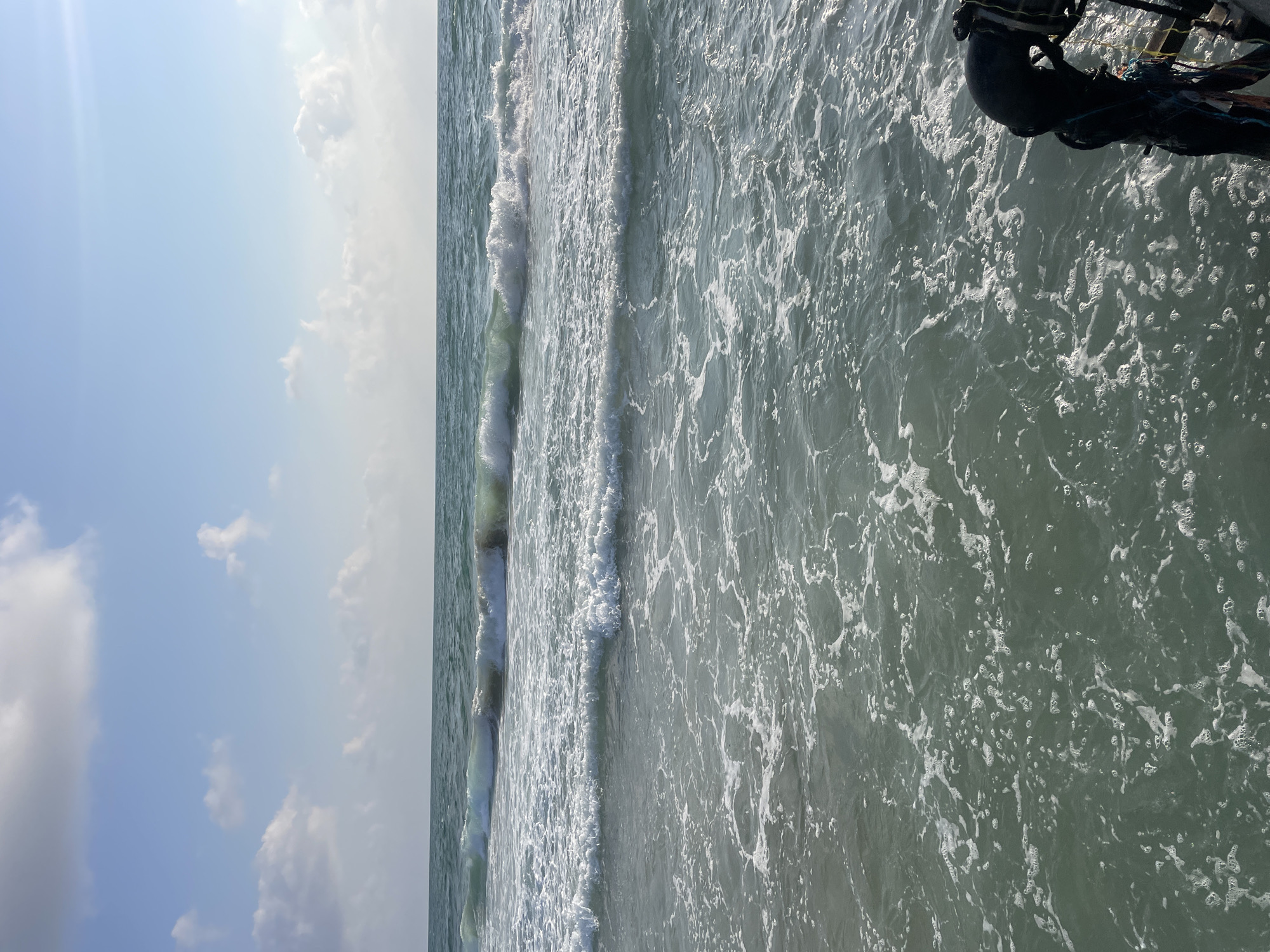 The image depicts a serene seascape under a partly cloudy sky, with waves gently rolling towards the shore. The water is a muted green, reflecting the sky and creating a peaceful atmosphere. In the foreground, a black object, possibly part of a pier or a boat, is visible on the right side, adorned with colorful ropes. The horizon is a sharp line separating the sea from the sky, with soft white clouds scattered across the blue expanse. This scene could be characteristic of the coastline in Haifa, Israel, known for its beautiful Mediterranean beaches and tranquil waters. The overall impression is one of calm and natural beauty.