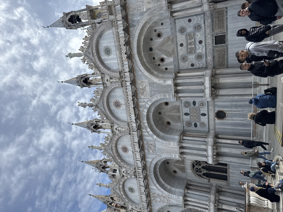 The image depicts the elaborate facade of St. Mark's Basilica (Basilica di San Marco) in Venice, Italy, viewed from a slightly low angle in St. Mark's Square. The sky is overcast with scattered clouds.
The basilica's lower and upper sections are visible, constructed primarily of grey and white marble with intricate details. The lower section features a series of grand arches, some serving as portals, with decorative columns and relief carvings. Ornate patterns and circular elements are embedded in the marble panels between these arches. A large, multi-paneled rose window is visible on the far left. The upper section showcases multiple pointed spires, adorned with statues and smaller architectural forms, rising against the cloudy sky. Pigeons are perched on several ledges and decorative elements of the building.
In the foreground and middle ground, approximately twenty individuals are present. They are dressed in casual, temperate weather clothing, including hoodies, jackets, and jeans. On the left, several people are seated on stone steps or a low wall: one person wears a blue beanie and black coat, another a black top, and a third a light top. A metal barrier or low railing extends across the middle ground. To the right of this barrier, a group of young men stands and interacts; one in a light grey hoodie holds a mobile phone, another wears a black hoodie, and a third a black puffer jacket. Other individuals are seen walking or standing throughout the scene. Backpacks are visible near some seated individuals. No overt interactions are visible between distinct groups, but people are in close proximity within the public square.