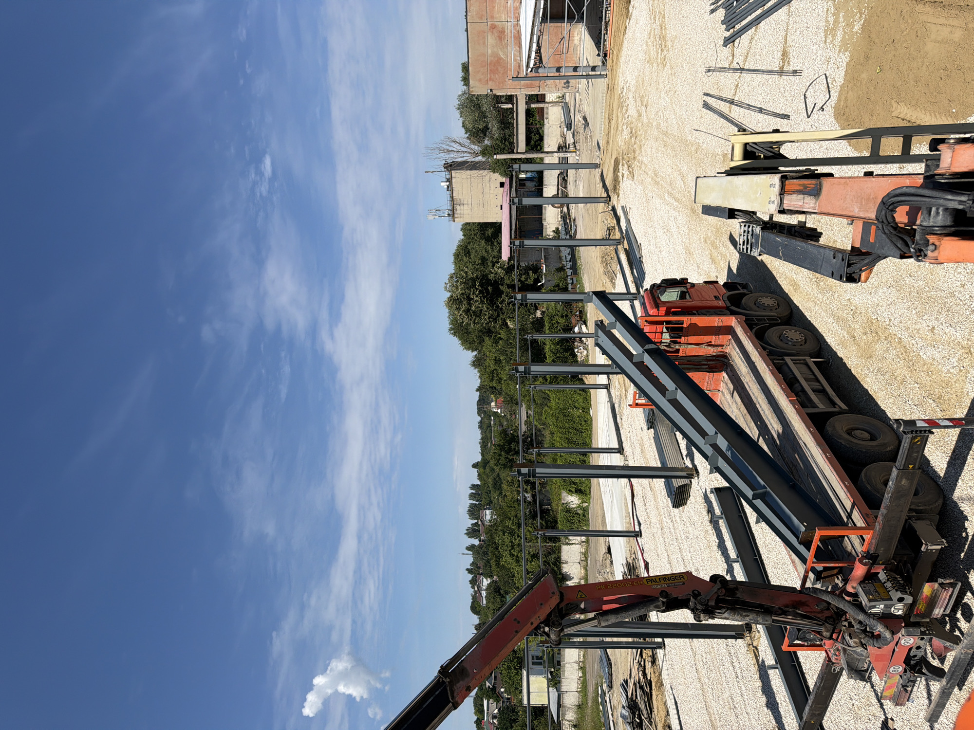An aerial, wide-angle shot captures an outdoor construction site on a clear, sunny day. The foreground is dominated by a red flatbed truck with an attached crane, its boom extended over a load of steel beams. A second, orange mechanical arm is visible on the right side of the truck. The ground is covered in light-colored gravel, with stacks of steel beams and rebar scattered throughout the scene. In the middle ground, several tall, dark grey steel columns stand upright, forming the skeleton of a new structure. Beyond the construction area, a line of green trees separates the site from a residential area with houses. In the background, a tall, concrete building with antennas on top is visible, along with other structures and the sky. The location is identified as Vatra, Moldova.