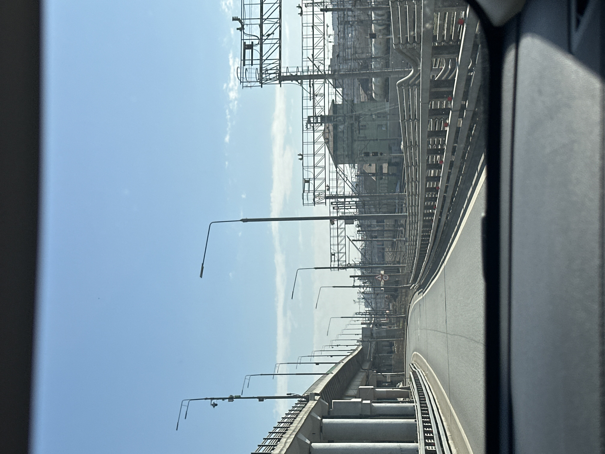 The image appears to be taken from inside a moving vehicle, looking out at a road infrastructure scene. The setting is likely an elevated highway or overpass, suggested by the rows of lampposts and the concrete supports visible beneath the road surface. On the right side of the image, heavy industrial structures with scaffolding and piping can be seen. The image shows Moscow, Russia.
