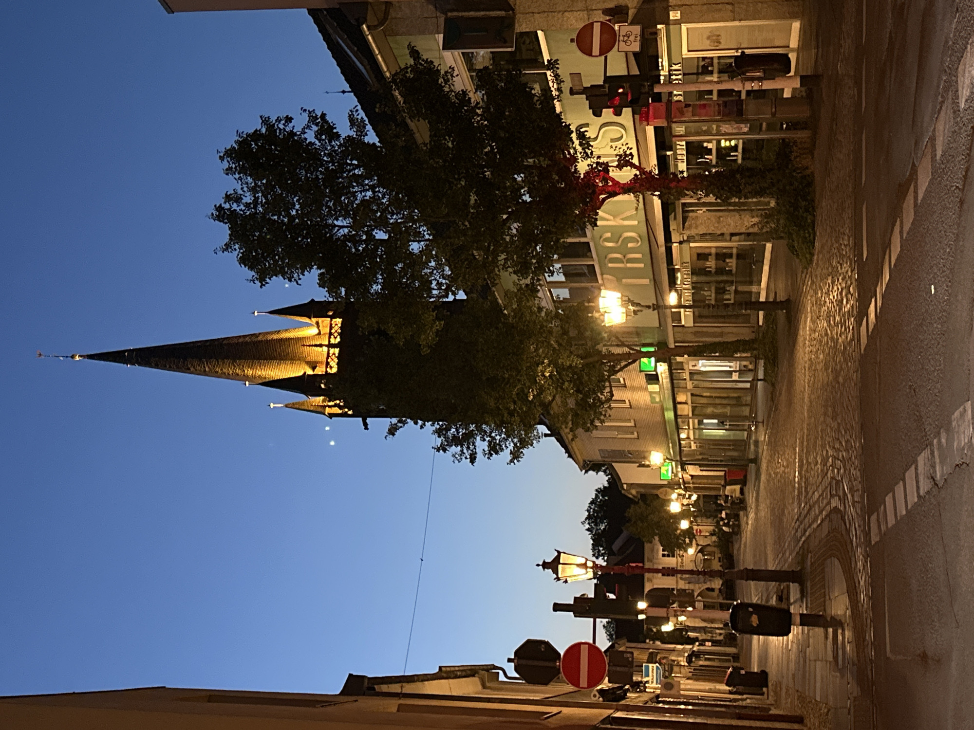 The image shows a street scene at dusk in Krefeld, Germany. A prominent church tower with illuminated spires dominates the background. The street is paved with cobblestones and lined with buildings that appear to be shops, some with illuminated windows. There are streetlights casting a warm glow on the street. Several traffic signs, including a "no entry" sign and a traffic light, are visible. There are trees along the street, some of which are illuminated by red lights. A crosswalk is marked on the street. In the distance, a few figures can be seen sitting at an outdoor cafe. The sky is a deep blue, indicating the transition from day to night. The overall impression is a quiet and atmospheric evening in a European city.