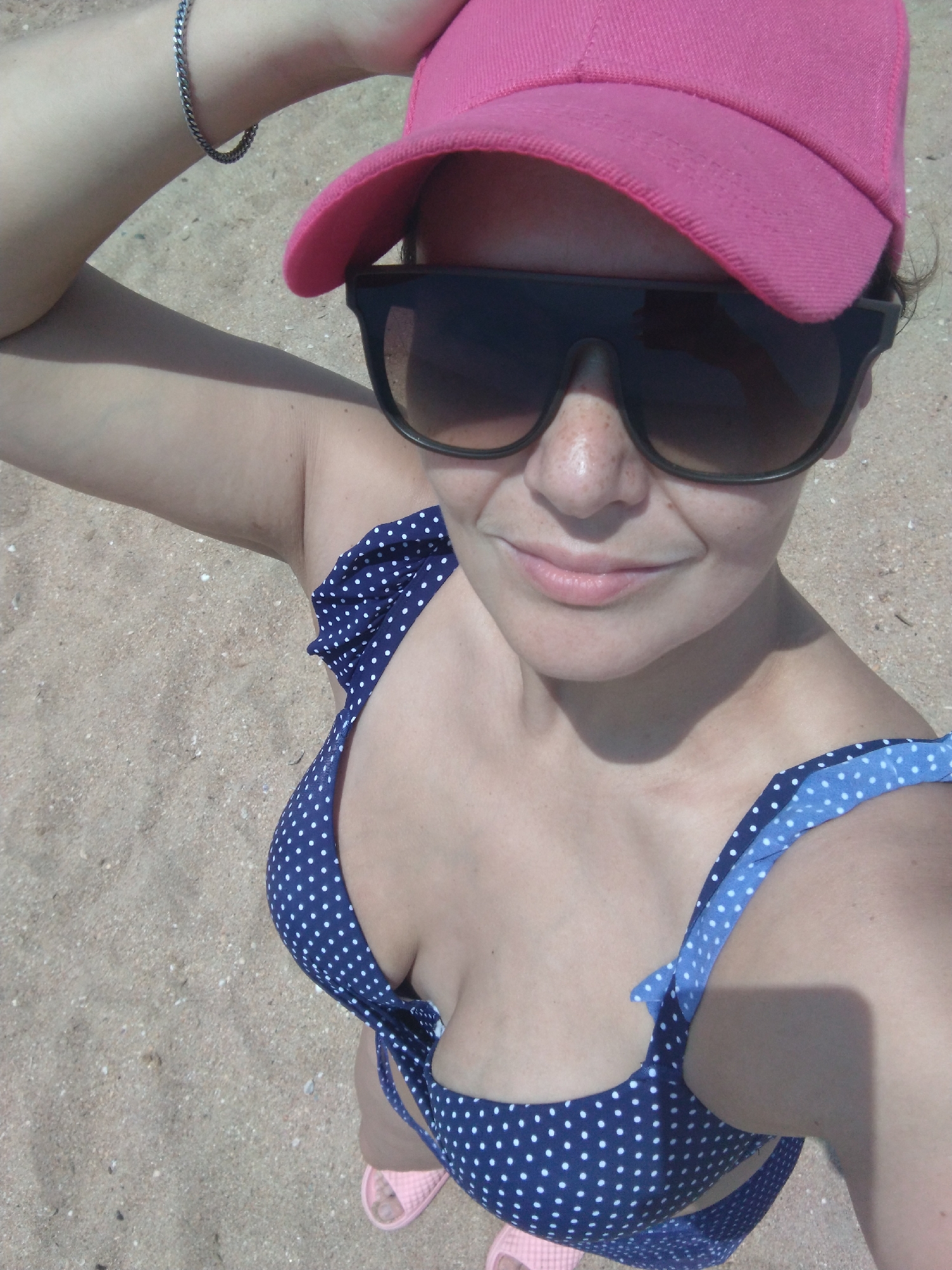 The image shows a close-up, high-angle, point-of-view shot of a person at a sandy beach, likely Vinogradny, Russia. The person is wearing a pink baseball cap, large dark sunglasses, and a blue and white polka dot bikini. They are holding their arm up, resting their hand on the brim of their cap. The sand is the dominant element of the background. The lighting suggests it is daytime, possibly a sunny day, with shadows visible on the person's arm and chest. The person appears to be smiling, indicating a relaxed and positive mood. No visible text is present in the image. The focus is on the person's face, upper body, and the beach setting.