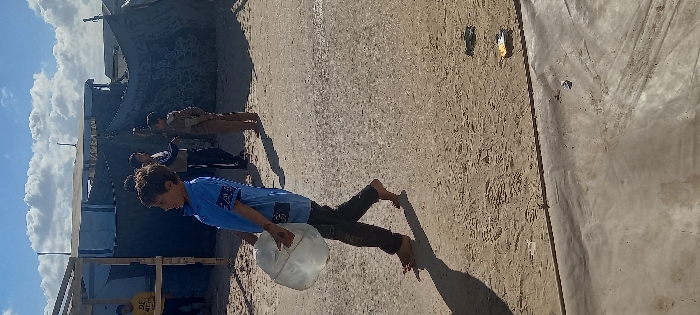 A young male, approximately 8-12 years old, walks barefoot from left to right across a sandy and dirt ground surface under a partly cloudy sky in Khan Yunis, Palestinian Territory. He wears a light blue short-sleeved shirt with dark markings and dark pants, carrying a clear, full plastic water container by its handle in his left hand. His shadow is cast long on the ground to his right. In the middle ground, several other males, appearing to be children or adolescents, stand near improvised structures. These structures include a wooden frame on the left, a dark tarpaulin tent with a light-colored top, and other rudimentary shelters. One individual in the background wears a yellow long-sleeved shirt. The ground also exhibits scattered debris, including plastic packaging. The scene is illuminated by strong sunlight, indicating an outdoor, daytime setting.