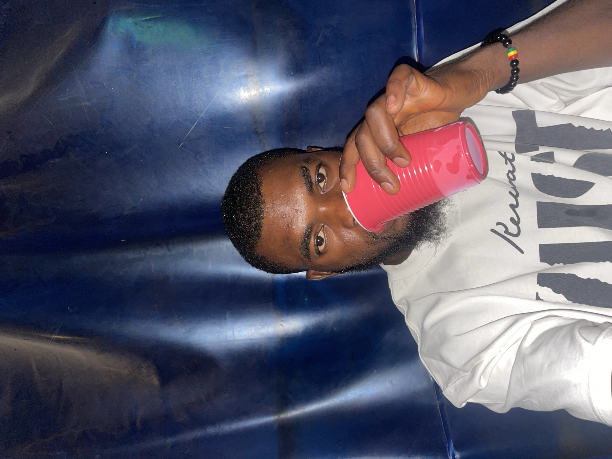 A medium shot, rotated counterclockwise, depicts a dark-skinned man with a trimmed beard, holding a red plastic cup to his lips, presumably drinking. The man is wearing a white t-shirt with black writing. He also has a beaded bracelet on his wrist. The background features a dark blue, glossy surface, possibly a wall or furniture. The location is not identifiable as Jos, Nigeria, based solely on the image.