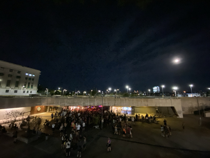 An outdoor night scene reveals a gathering of people under a bridge-like structure with colorful lights. The crowd is densely packed towards the center, thinning out towards the sides where some individuals are seated. Above, a row of streetlights illuminates the background and a bright moon shines in the dark sky. A large building is visible on the left side of the frame.