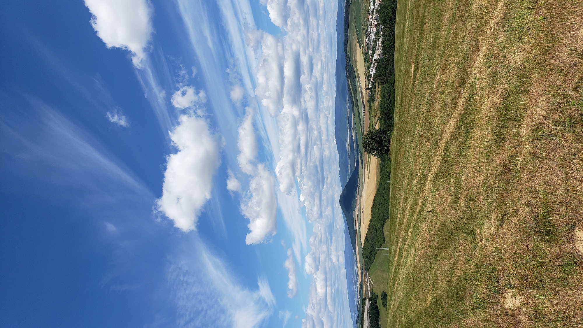 A rural landscape under a bright blue sky with scattered white clouds. In the foreground, a field of dry, mown grass shows parallel lines from a mower. Beyond the field, rolling hills are covered in green and yellow vegetation. A distinctive conical hill stands in the mid-ground, casting a shadow. A small town with buildings and trees is visible in the distance, nestled among the hills. The image appears to be taken on a sunny day. The overall scene suggests a tranquil countryside setting near the city of Prešov, Slovakia.