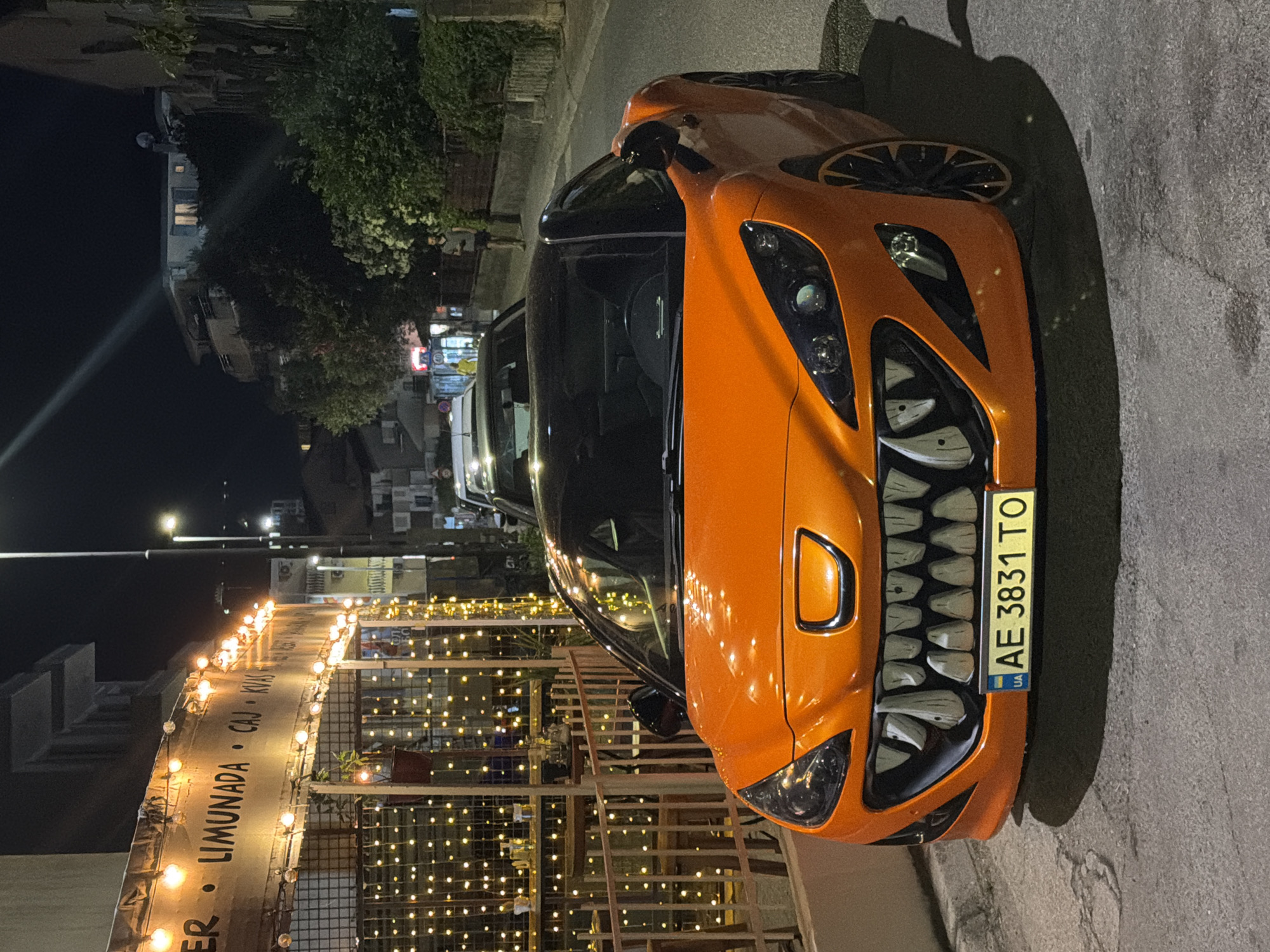 An orange sports car with a monstrous, toothy grille is parked on a paved street at night, in front of a restaurant or cafe decorated with numerous strings of warm-toned fairy lights. The car's headlights are on, illuminating the road. Behind the car, a building with a sign advertising "LIMUNADA," "CAJ," and "KVAS" is visible. To the right of the street, trees and buildings are dimly lit, suggesting a residential or mixed-use area within Budva, Montenegro. A streetlight casts a beam of light across the scene from the upper left. The overall atmosphere is nocturnal and slightly whimsical due to the car's unusual modification.