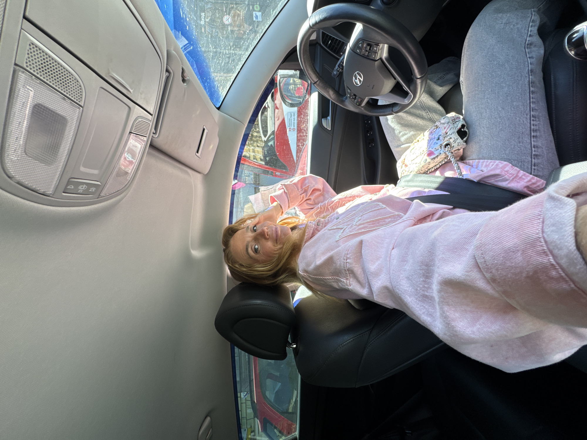 A woman wearing a pink jacket is seated in the driver's seat of a Hyundai vehicle. She is positioned in front of the steering wheel, wearing jeans and a white purse is on her lap. The vehicle is parked outside. The location is Saint Petersburg, Russia based on text visible on the nearby vehicles.