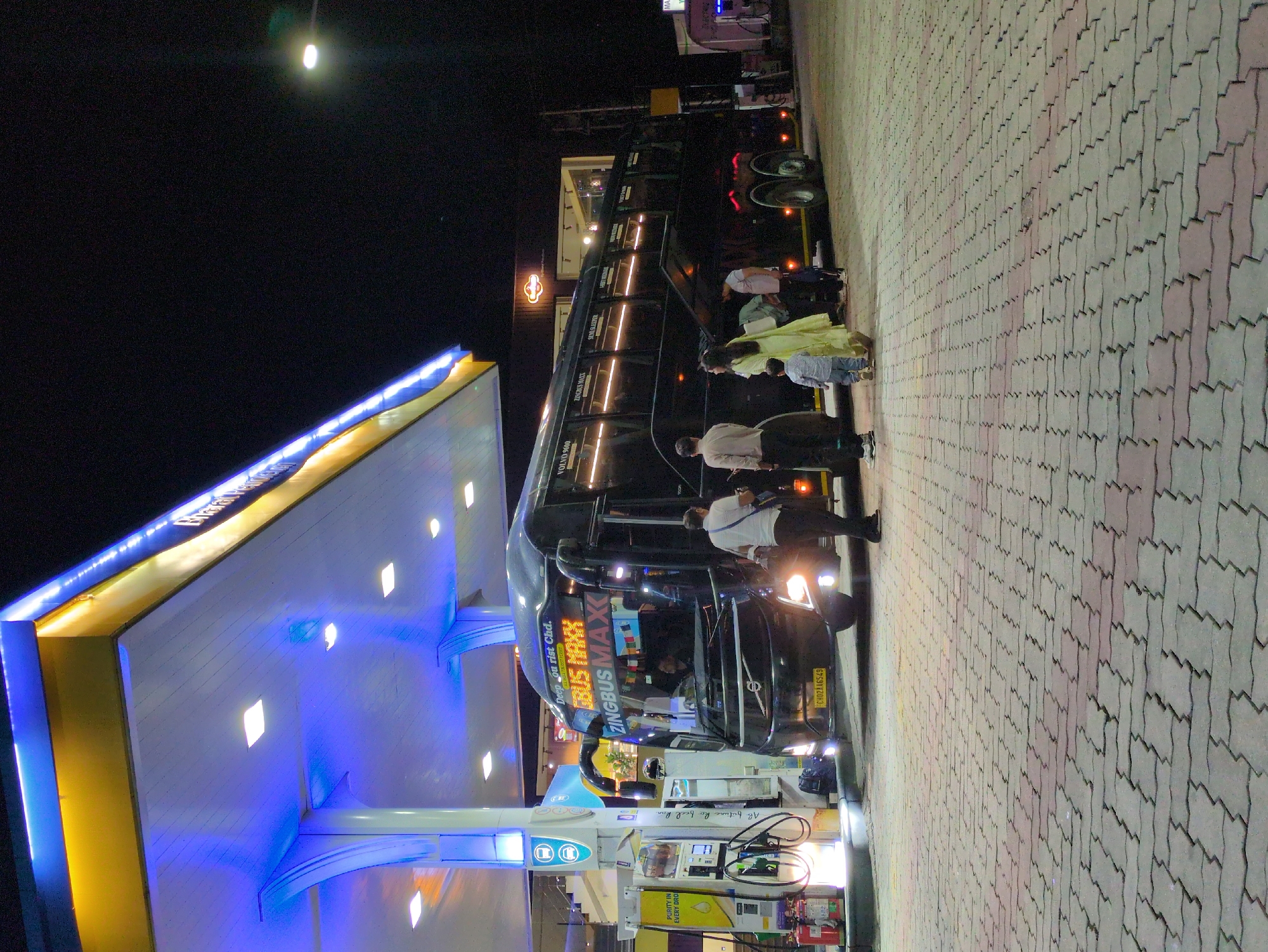 This is a low-angle, night-time photograph taken at a Bharat Petroleum petrol station in Dharamshala, India. The primary subject is a black Volvo 9600 bus with "ZINGBUS MAXX" displayed on its front. Several people are gathered around the bus. A man in a white shirt and dark pants stands to the right of the bus entrance, and a woman in a light yellow dress with a child in a blue shirt and grey pants are walking towards the bus on the right side. Another man in a light brown shirt and dark pants is also near the bus. The petrol station canopy is illuminated with blue and yellow lights. There are fuel pumps visible to the left of the bus, along with fire extinguishers and a backpack. The ground is paved with interlocking bricks. The overall atmosphere is that of a public transportation stop at night.