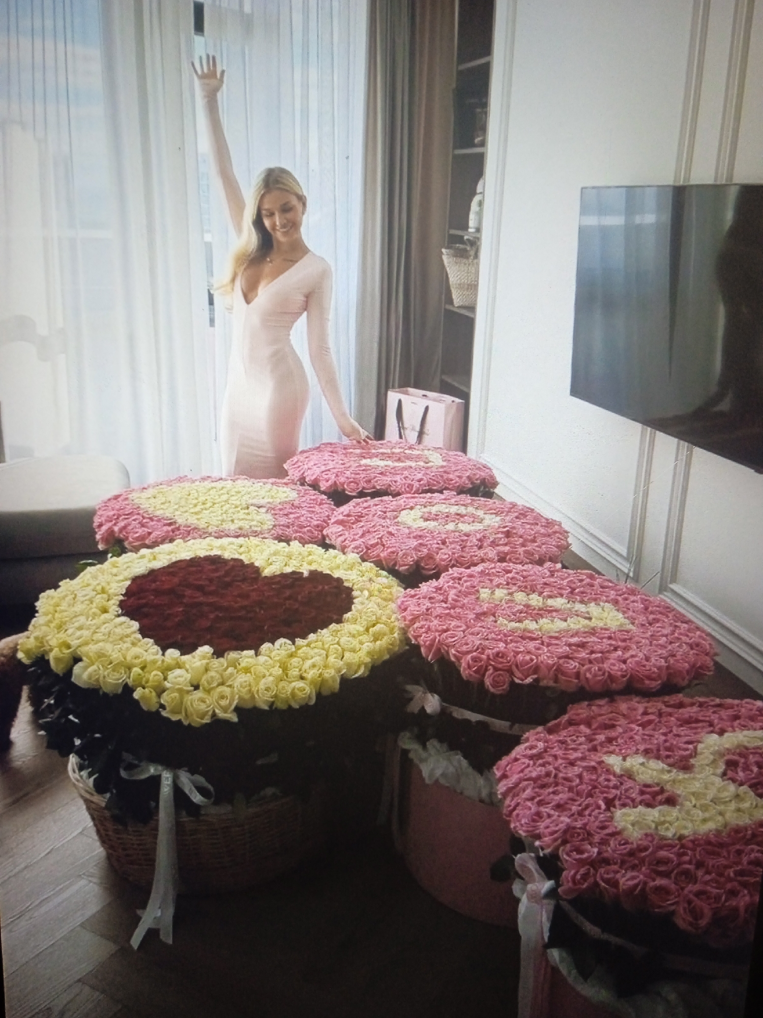 A blonde woman in a form-fitting, long-sleeved pink dress stands in a well-lit room with a large window and sheer curtains. She has her left arm raised and is smiling. In front of her, arranged on a wooden floor and on a dark surface, are several large, heart-shaped floral arrangements made of pink and yellow roses. One of the largest arrangements, in the foreground, is a heart shape outlined in yellow roses with a red rose center, placed in a woven basket with a white ribbon. Other arrangements are primarily pink roses with yellow accents forming heart shapes, some in baskets and some in pink boxes. In the background, a bookshelf with various items, a pink bag, and a television are visible. The scene suggests a luxurious indoor setting. The location is not specified in the image, and therefore cannot be determined to be Chernivtsi, Ukraine.