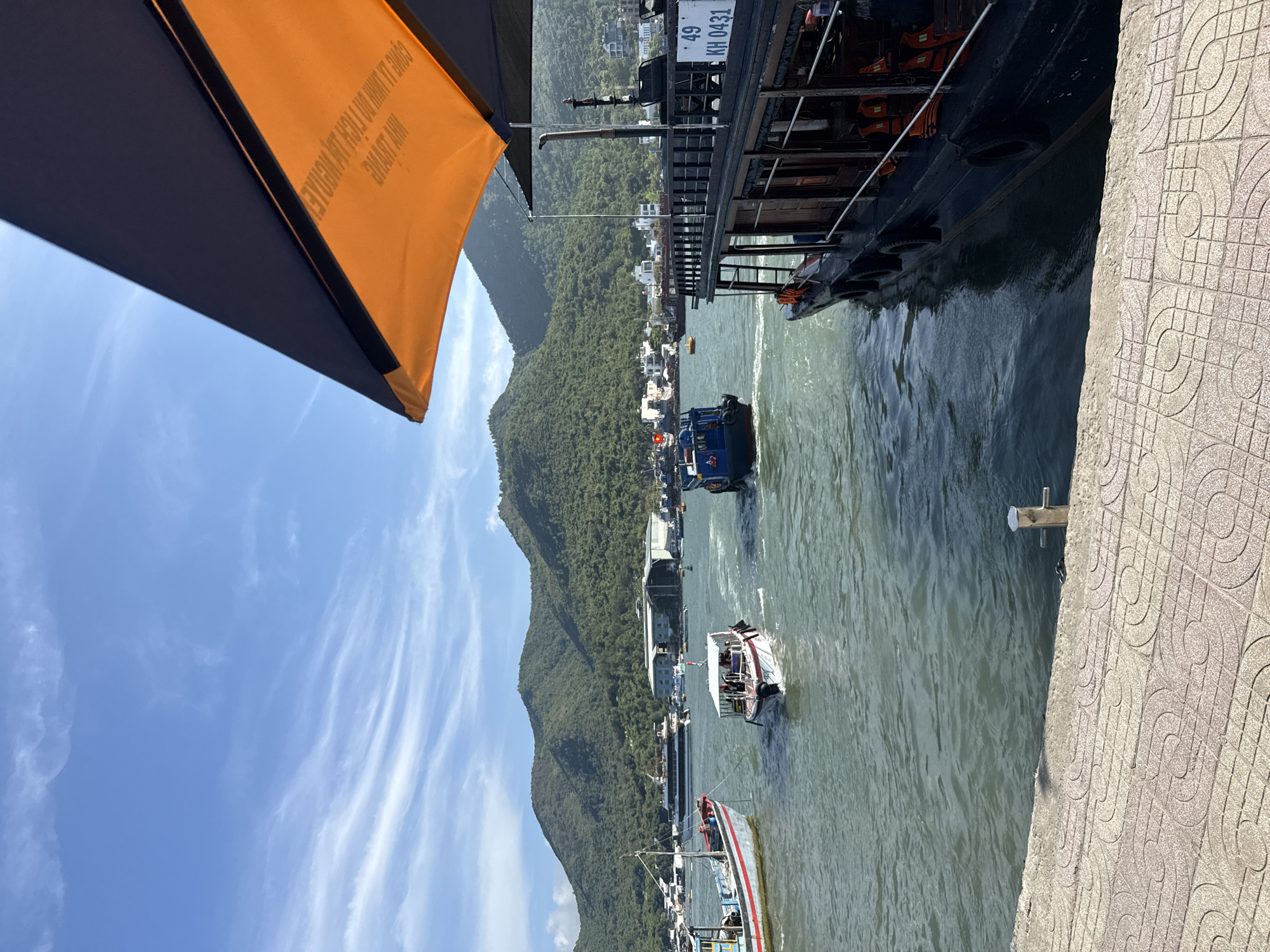 The image captures a view of a harbor or bay in Nam Nha Trang Ward, Vietnam. Dominating the frame on the right is the side of a large boat or ferry, identifiable by the number "49 KH 0431" displayed on its side. Life preservers are attached to the boat's exterior. The water is visible, with several smaller boats navigating through it. In the background, a lush green mountain range rises from the shoreline. A section of a city or town with buildings is nestled along the coast at the base of the mountains. The sky above is blue with scattered white clouds. On the left side of the image, a portion of a parasol or awning is present, with some text in a non-English language. The perspective suggests the photograph was taken from a pier or embankment, with a patterned concrete surface visible in the lower right corner.