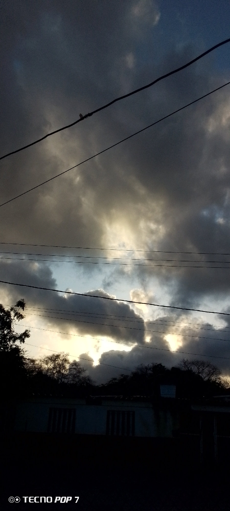 The image shows a cloudy sky with electrical wires running across it. The sun is trying to peek through the clouds, casting a warm light. Below the sky, there is a silhouette of trees and a building. The location is in Altagracia, Venezuela.