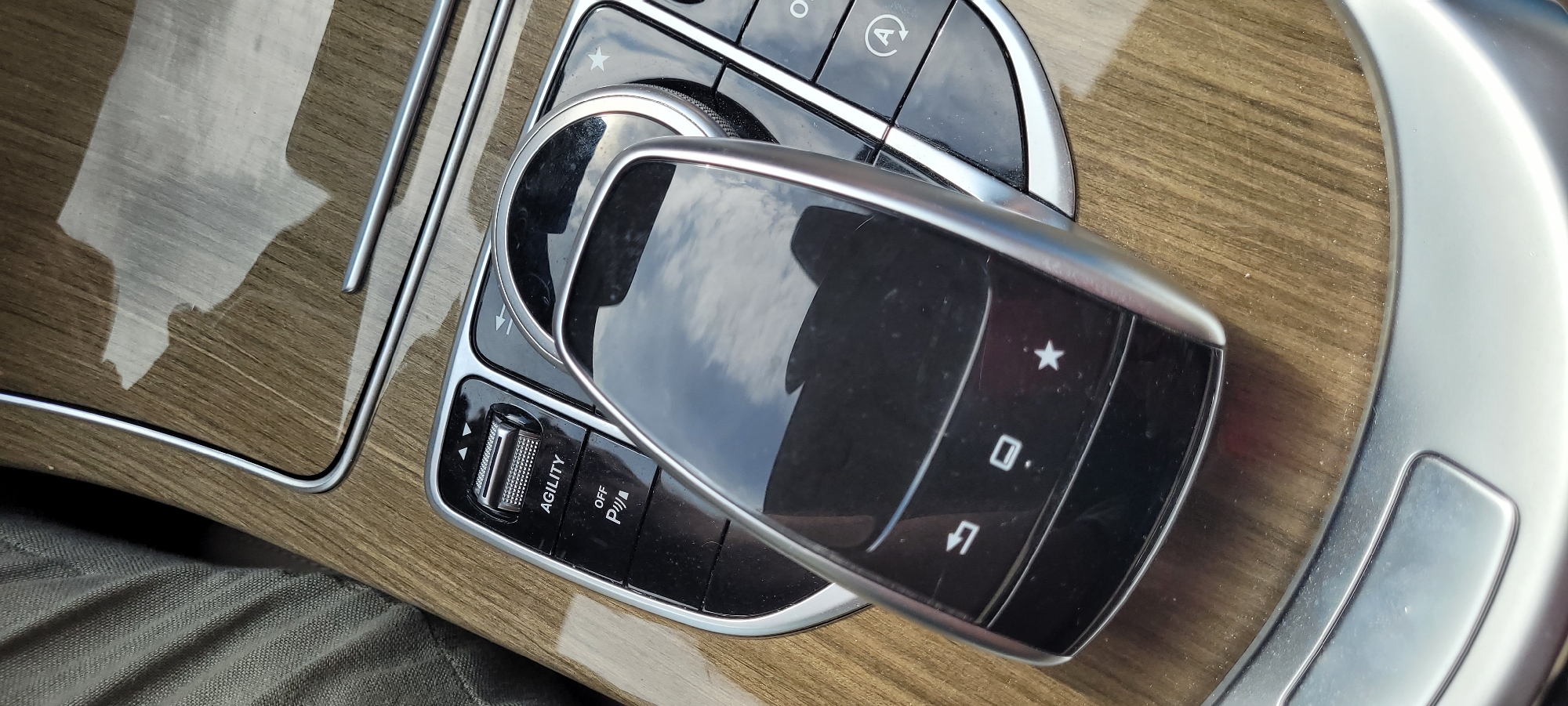 This is a close-up, eye-level, vertical shot of a modern car's center console. The console features wood grain trim with a high-gloss finish. Dominating the foreground is a black, reflective car key fob, possibly a Mercedes-Benz key, with a silver metallic accent along its edge. The fob is angled, lying on top of a rotary dial control.
The rotary dial is surrounded by a chrome bezel and sits within a cluster of black buttons. To the left of the rotary dial, there is a rectangular button with the word "AGILITY" printed on it, along with directional arrows above and below. Below the "AGILITY" button are two more buttons, one labeled "OFF" with a parking sensor icon (P and a car outline) and another with a circled "A" icon, likely for the start-stop system.
The background consists of the wood-grained console and a hint of a person's knee or leg covered by grey, textured fabric to the left. The lighting suggests an indoor or shaded environment, possibly a car interior, with reflections of the sky visible on the shiny surfaces of the key fob and console.
This scene is not specific to Riga, Latvia, or any other city, as it depicts the interior of a vehicle and lacks any identifiable external landmarks or urban context.