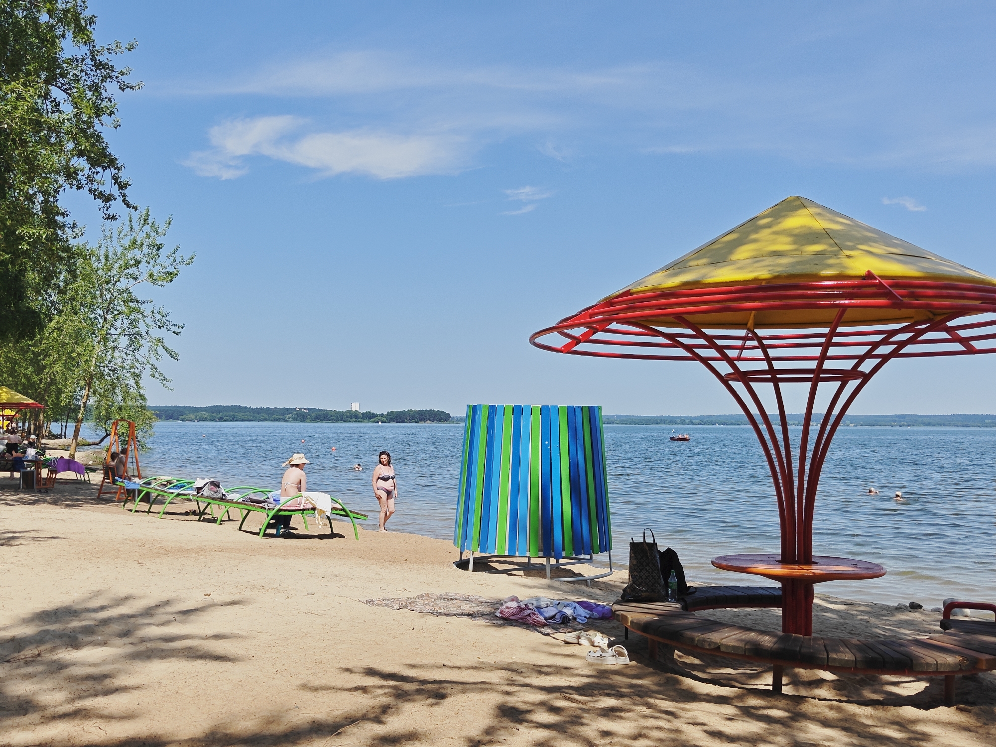 A sunny day at a lake beach in Belarus, likely during the summer. The scene depicts a sandy beach with several people enjoying the water and relaxing.
In the foreground, a large, decorative red and yellow umbrella structure with a circular wooden bench beneath it stands prominently. Beach chairs are set up on the sand, some occupied. A striped changing booth, featuring vertical blue and green panels, is positioned near the water's edge.
Further back, people are visible in the shallow water, swimming and enjoying the lake. The water is calm and reflects the clear blue sky, which has a few wispy clouds. Trees line the left side of the beach, casting shadows on the sand, suggesting it's mid-day. The overall atmosphere is one of leisure and recreation.