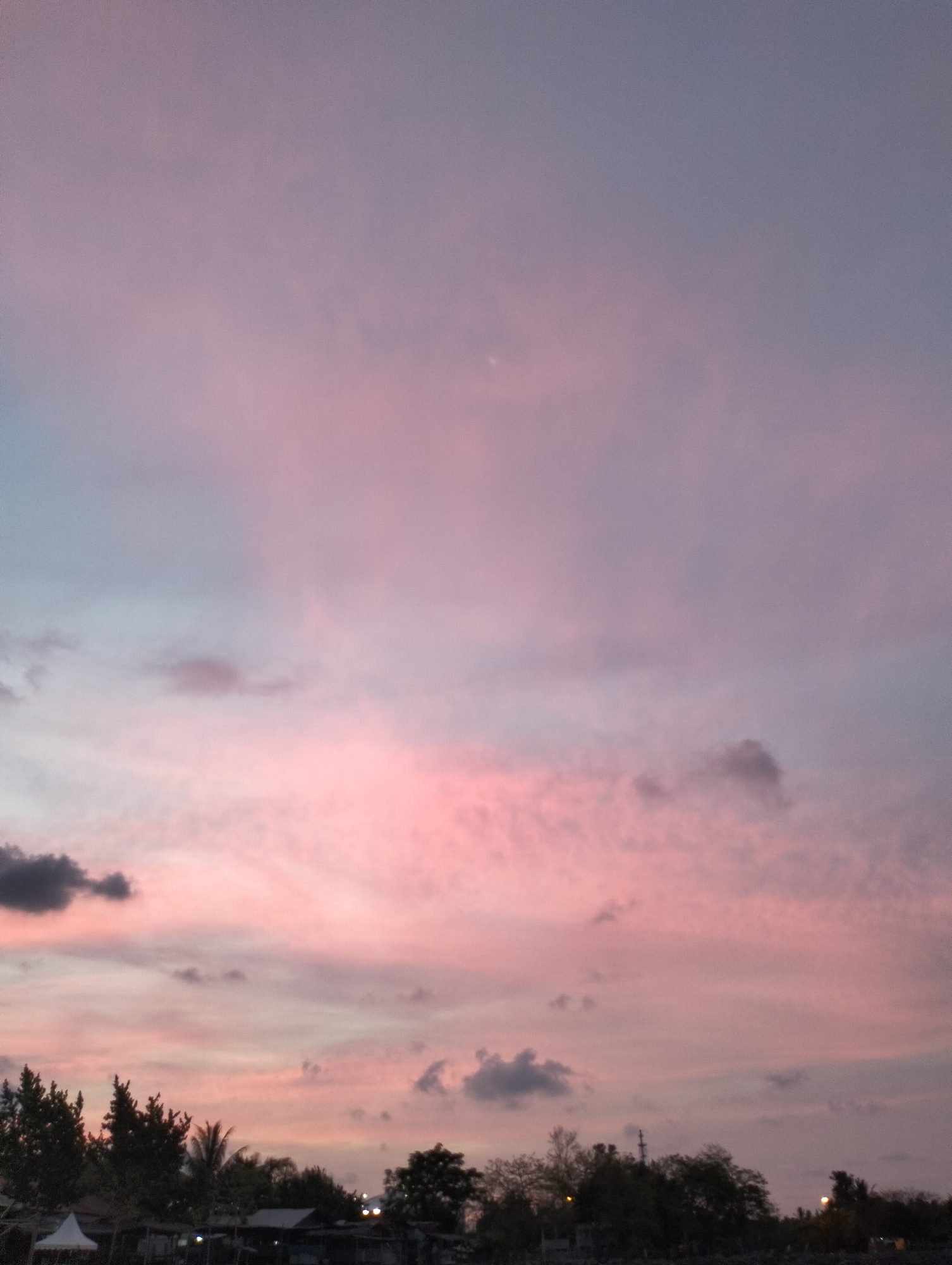 This image captures a wide shot of a twilight sky over Lhokseumawe, Indonesia, dominated by expansive clouds painted in various shades of pink, purple, and blue-grey. The time of day is likely sunset or sunrise, given the soft, warm illumination coloring the clouds. The upper part of the sky transitions from a lighter pink-purple at the horizon to a deeper blue-grey towards the top.
In the lower section of the frame, the silhouettes of numerous trees and dense tropical foliage form a dark, irregular horizontal band across the scene. Below the tree line, indistinct buildings and structures are visible as dark shapes, with a white tent or awning faintly discernible on the far left. Scattered among these darkened structures, several small, bright points of light suggest artificial illumination from the ground. There are no identifiable people or specific activities taking place. The atmosphere conveyed is serene and peaceful, emphasizing the natural beauty of the sky at dusk or dawn. No visible text is present in the image.