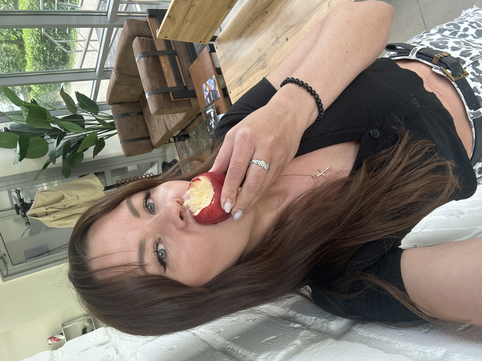 A woman with long brown hair, wearing a black top and leopard-print shorts, takes a selfie while eating an apple. She is indoors, near a large window with a view of greenery. The room appears to be a lounge area with a brown couch, wooden furniture, and a large potted plant. This location is in the city of Chaikovsky, Russia.