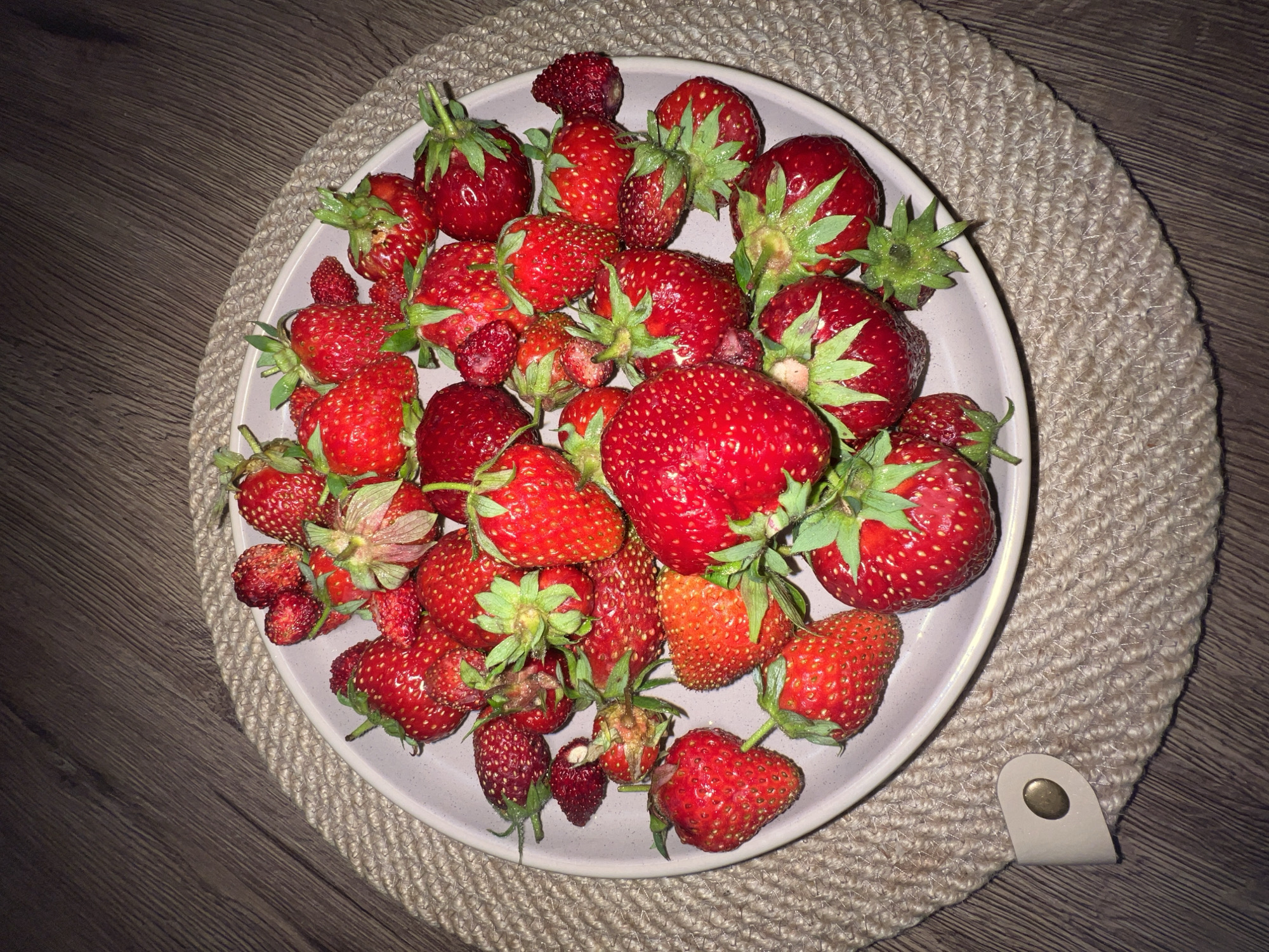 A top-down, medium shot captures a white plate filled with ripe, red strawberries. The plate is placed on a round, woven placemat, which rests on a wooden surface. The strawberries are varied in size and shape, with some appearing plump and smooth, while others are smaller and more textured. Green leaves and stems are visible on many of the fruits. The lighting creates subtle shadows, highlighting the textures of the strawberries and the woven placemat. The wooden surface shows a natural grain pattern. No people or animals are present in the image. The scene appears to be indoors, possibly in a kitchen or dining area, given the tableware and wooden surface. The context does not provide specific information about the location within Votkinsk, Russia, beyond the fact that the image was taken there.