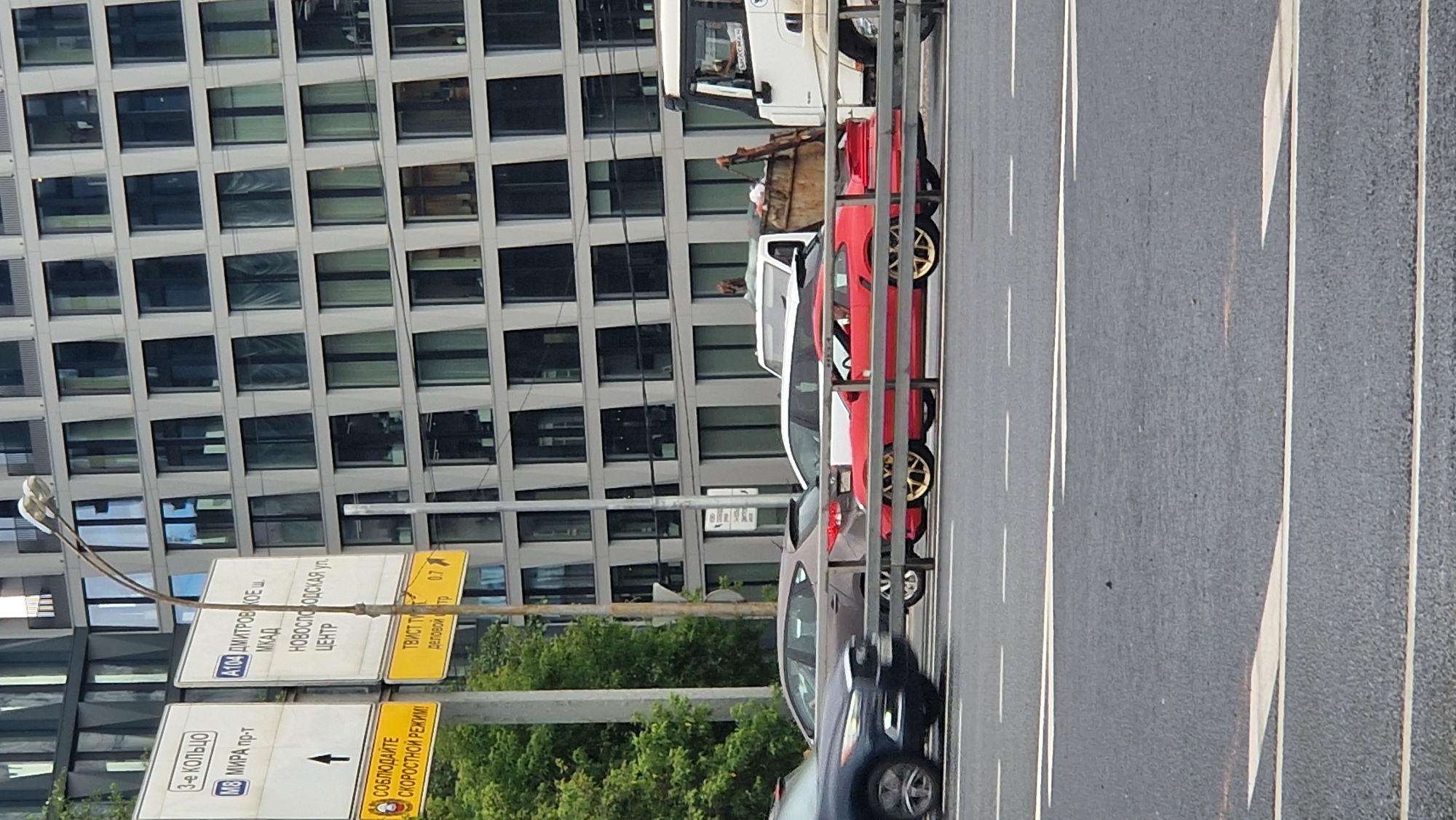 The image shows a street scene in Moscow, Russia. The foreground is dominated by a multi-lane road with white markings. Several cars are parked or driving along the right side of the road. Notably, a red sports car with gold rims is visible, alongside other vehicles. In the background, a modern, grey building with a grid of windows rises prominently. The time of day appears to be daytime, with the lighting suggesting it might be late afternoon or early evening. The weather seems to be overcast, as there are no harsh shadows and the sky is not clearly visible.
Several signs are present, providing directional and informational cues. One sign indicates "3-е КОЛЬЦО" (3rd Ring Road) and "МИРА пр-т" (Mira Avenue) with an arrow pointing forward. Another sign displays "4109 ДМИТРОВСКОЕ Ш." (4109 Dmitrovskoe Highway), "МКАД" (MKAD - Moscow Ring Road), and "НОВОСЛОБОДСКАЯ УЛ. ЦЕНТР" (Novoslobodskaya Street, Center), along with the distance "0.7". A yellow sign reads "СОБЛЮДАЙТЕ СКОРОСТНОЙ РЕЖИМ!" (Observe the speed limit!). Further text on a yellow sign appears to be "ТВИСТ ТУ" and "деловой тр". A white truck with the brand "SHACMAN" is also visible, possibly a garbage truck given the container behind its cabin. The overall impression is a busy urban environment with traffic and modern architecture.