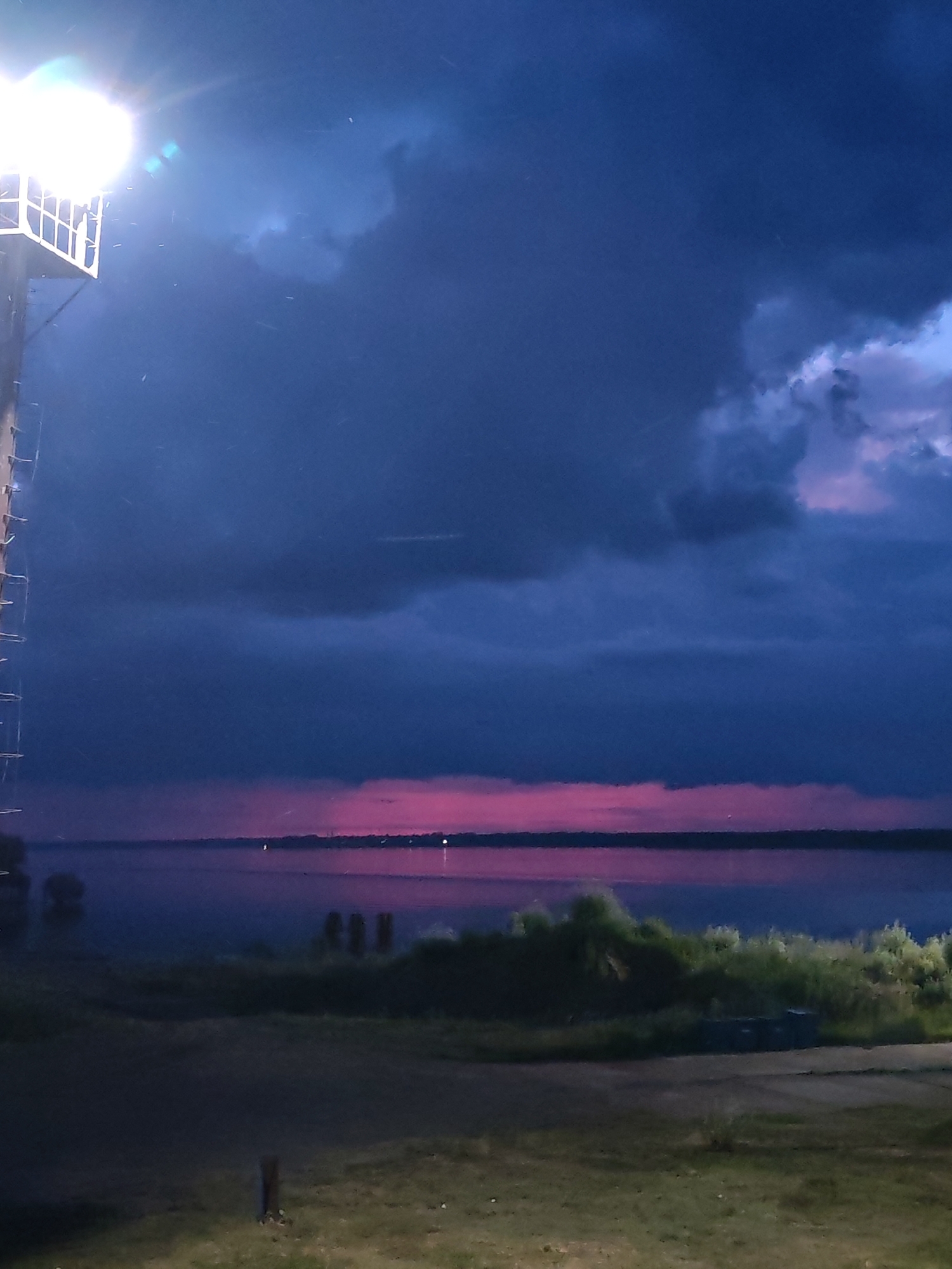 The image depicts a dramatic twilight scene over a body of water, likely a river or lake, in Rybinsk, Russia. Dominating the sky are large, dark, ominous storm clouds, tinged with deep blues and purples. A striking band of vibrant pink and magenta hues cuts across the horizon, suggesting the last rays of sunset or the prelude to a storm.
In the foreground, a large, white, industrial-looking light fixture, possibly part of a tower or structure, illuminates the scene with a bright, cool-toned light. Tiny particles, possibly rain or dust, are visible suspended in the air, caught in the beam of the light. Below the light, a metal ladder or railing descends along the side of the structure.
The middle ground features a stretch of dark shoreline, with silhouetted vegetation, including what appear to be bushes and trees, casting shadows. In the water, a few indistinct shapes suggest the presence of people or small boats, their forms barely discernible against the dark water. Further out on the water, faint pinpricks of light suggest distant settlements or lights. The overall mood is somber and atmospheric, with the dramatic sky and approaching darkness creating a sense of foreboding or awe. The time of day is clearly evening, during or just before a storm. No visible text is present in the image.