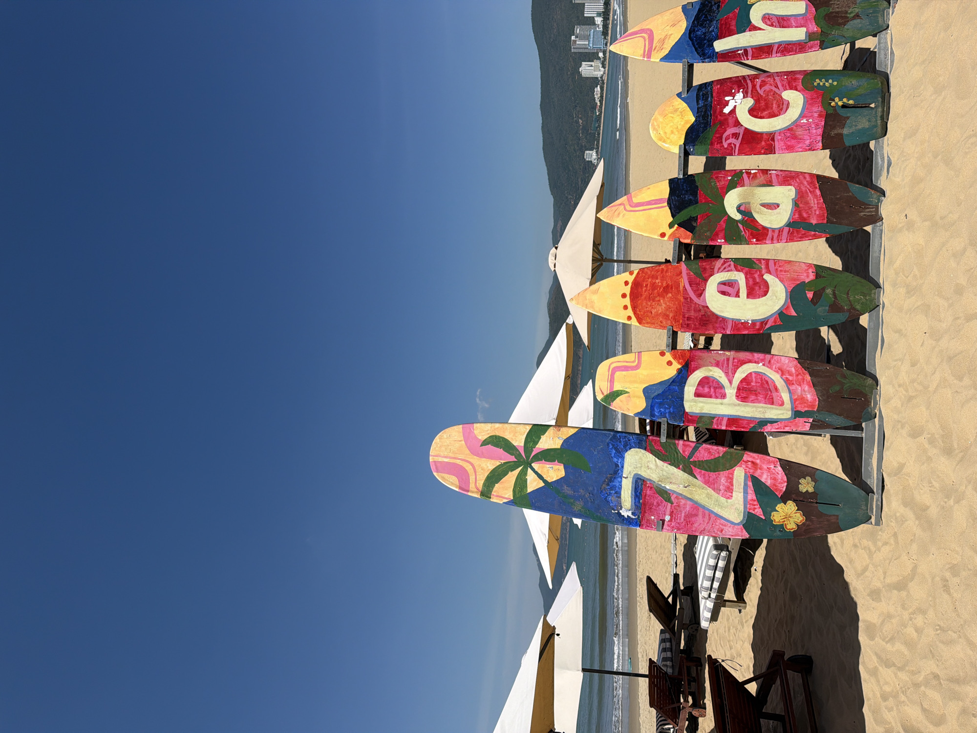 This is a vertical, eye-level, medium shot photograph of a beach scene in Quy Nhon Ward, Vietnam. In the foreground, a row of decorative surfboards stands upright on the sand, spelling out "ZBeach." Each surfboard is painted with vibrant colors and tropical motifs, including palm trees, mountains, and flowers. The surfboards are arranged from left to right, with the first one displaying a stylized "Z" and the subsequent ones displaying "B," "e," "a," "c," and "h."
Behind the surfboards, the sandy beach stretches towards the ocean. Several beach chairs and umbrellas are visible, indicating a recreational area. The ocean is calm with gentle waves. In the background, a coastline with a city skyline featuring buildings and mountains can be seen under a clear blue sky. The overall scene depicts a sunny day at a tropical beach destination. There are no people visible in the image.