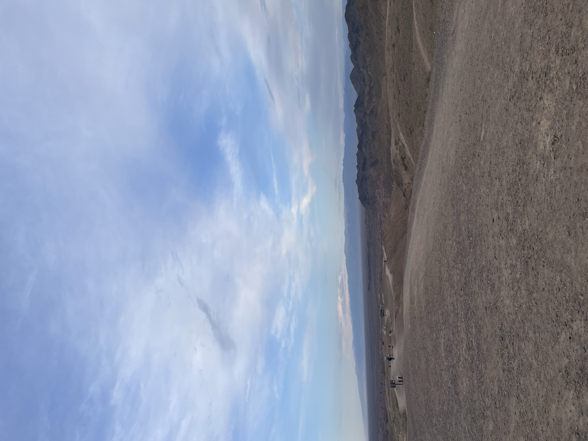 The image depicts a vast, arid landscape with rolling hills and distant mountains under a partially cloudy sky. A dirt path winds through the foreground, leading towards a group of people scattered across a plateau. Several individuals are walking on the path, and there are structures that appear to be informational signs or resting benches. The terrain is characterized by dry, reddish-brown earth, with sparse vegetation. The background reveals layers of eroded hills and a long, flat horizon line where the earth meets the sky, with a mountain range visible in the far distance. The sky itself is a mix of pale blue and white clouds, with some darker streaks indicating potential weather patterns. This scene is characteristic of the Charyn Canyon National Park, located near Tamerlik in Kazakhstan, known for its unique geological formations and panoramic views.