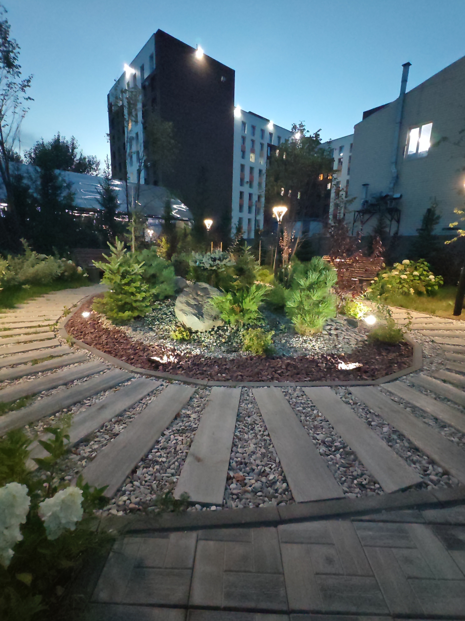 In Kokshetau, Kazakhstan, an exterior shot captures a landscaped garden area at dusk. The scene is dominated by a central, circular garden bed featuring a large boulder, various green shrubs, and small evergreen trees. The garden is illuminated by strategically placed spotlights, highlighting the textures of the stones and foliage. A curved pathway, constructed with alternating wooden planks and gravel, leads into the garden. In the background, modern apartment buildings with illuminated windows are visible against the twilight sky, suggesting a residential or urban setting. The overall atmosphere is serene and well-maintained, indicating a thoughtfully designed public or private space.