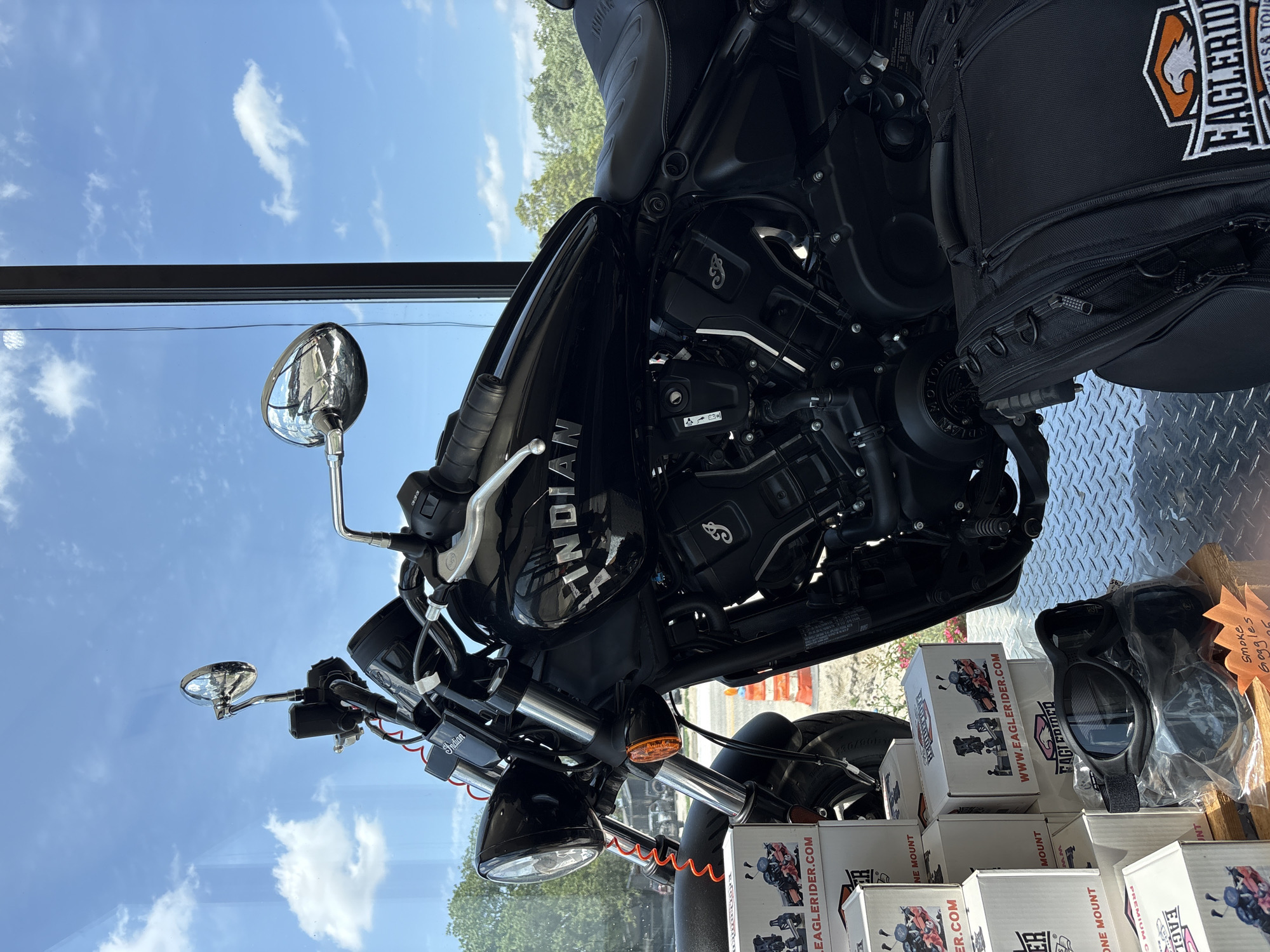 A black Indian motorcycle is displayed in a showroom, likely at Eagle Rider, a motorcycle rental and tour company, given the branding on the boxes and bags. The motorcycle is parked indoors, with a large window in the background revealing a bright blue sky with scattered white clouds, and trees and buildings visible outside. Several cardboard boxes, some with branding and images of motorcycles, are stacked to the left of the motorcycle. A pair of black goggles sits on top of one of the boxes. A black motorcycle bag with the Eagle Rider logo is placed on the floor to the right of the motorcycle. The scene appears to be a sunny day, and the motorcycle is clean and in good condition, suggesting it is for sale or rent. The overall impression is of a well-lit and organized display aimed at motorcycle enthusiasts.