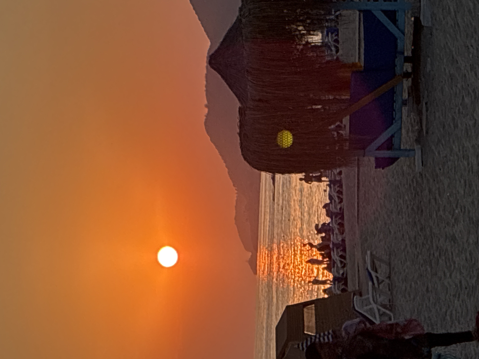 A vertical, medium shot captures a tranquil sunset scene at Kleopatra Beach in Alanya, Turkey. The sun, a bright, white orb, is low in the sky, casting a warm orange glow across the horizon and reflecting vibrantly on the calm sea. Silhouettes of people are visible enjoying the beach, some standing near the water's edge, others scattered among rows of empty lounge chairs.
In the foreground, a beach umbrella with a striped pattern stands to the left, partially obscuring a dark-colored structure. To the right, a thatched-roof beach hut with a wooden support structure is prominent. A yellow light fixture is visible through the thatched roof. Beyond the beach hut, more lounge chairs are arranged. In the distance, a chain of mountains forms a silhouetted backdrop against the sunset sky. A person in dark clothing is standing in the lower left quadrant of the image. The beach itself is composed of fine sand. The overall atmosphere is serene and highlights the beauty of a Mediterranean sunset.