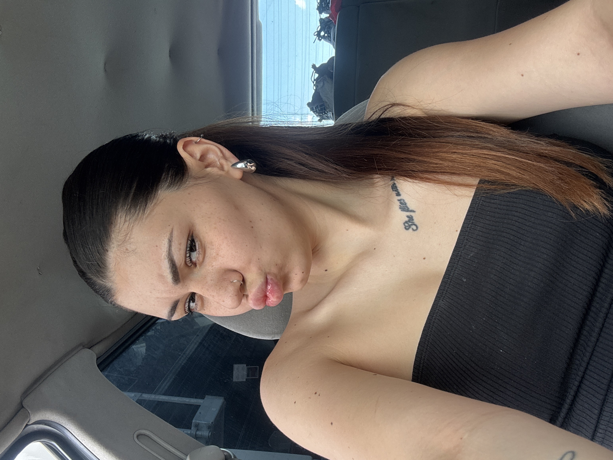 A young adult female is positioned in the foreground, seated within the passenger compartment of a vehicle. She faces the viewer directly, her lips pursed in a pout, and her eyebrows slightly furrowed. Her skin exhibits numerous freckles across the nose and cheeks. She has a silver hoop nose piercing in her left nostril and multiple earrings in her left ear, including a silver hoop in the upper cartilage and a larger, reflective silver teardrop-shaped earring in the lobe. Her hair is dark at the roots, transitioning to a lighter auburn or brown shade towards the ends, and appears to be partially pulled back, with some strands draped over her right shoulder. She wears a black, ribbed, strapless top. A tattoo with text starting "She flies with" is visible on her upper left chest.
The interior of the vehicle features grey upholstery on the seat and a grey headliner. Through the rear window, a light blue sky and indistinct distant structures or natural elements are visible. On the rear seat, several pairs of shoes, including sneakers, are present. A car door frame or pillar is visible on the left side of the image. There are no discernible landmarks, specific architectural features, or unique geographic indicators within the image to confirm the location as Sofia, Bulgaria, or any other particular city.