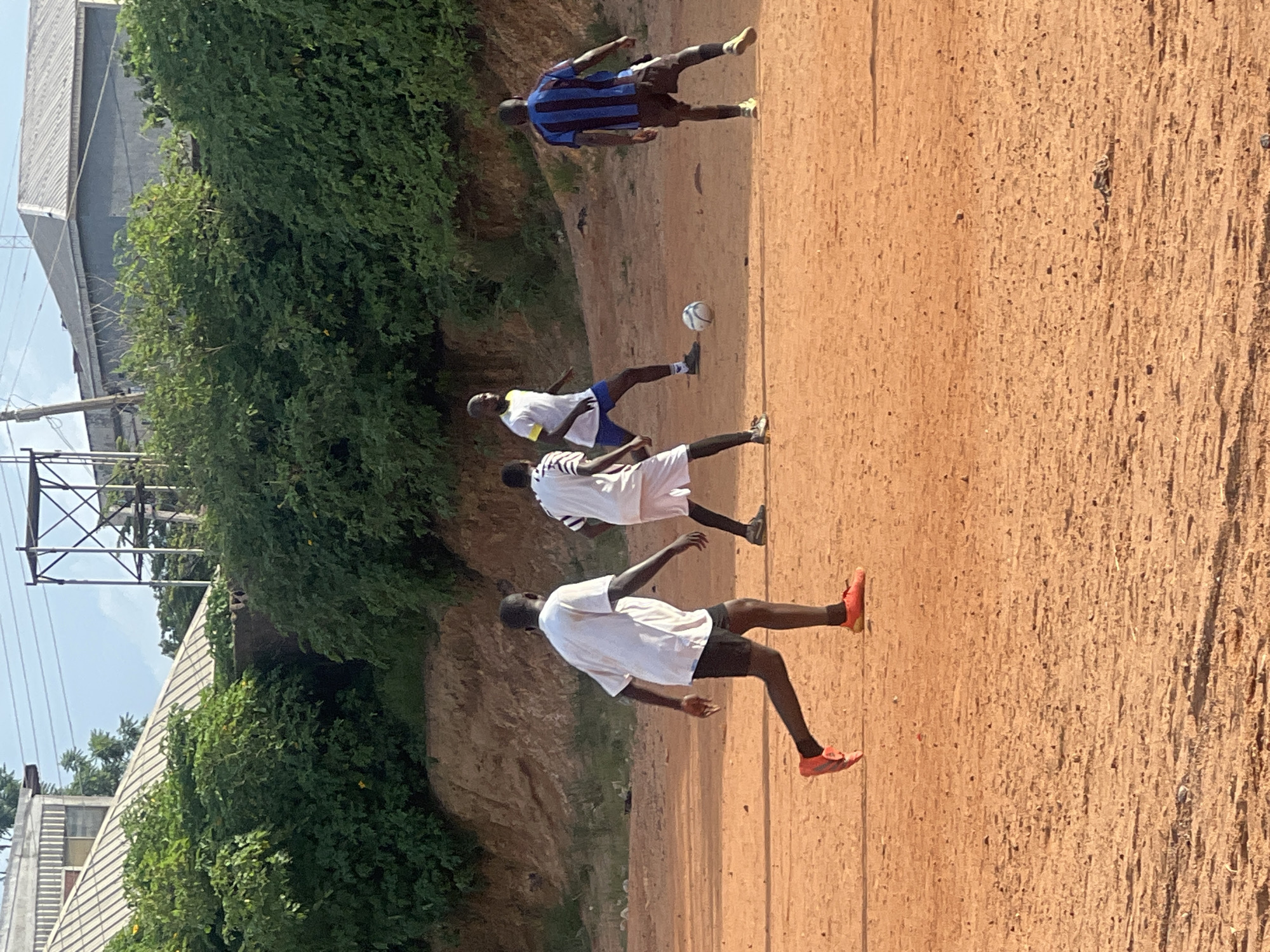 The image shows four young Black men playing soccer on a dirt field under a bright, overcast sky. The field is a reddish-brown color, with some scattered rocks and debris. In the background, there is a dense green hedge, a sandy embankment, and some buildings with corrugated metal roofs. There is also a tall, metal structure, possibly a water tower, visible against the sky. The men are all wearing athletic clothing. One man in a white shirt and black shorts is in the foreground, facing away from the camera, with his right leg extended and his left leg bent. Another man in a white shirt and white shorts is to his left, also facing away. A third man in a blue and black striped shirt and brown shorts is to the right, facing away from the camera and slightly towards the man in the foreground. A fourth man in a white shirt with yellow accents and blue shorts is in the background, facing towards the man in the foreground and looking at the soccer ball between them. The soccer ball is white and appears to be in motion. The overall impression is of a casual pickup soccer game. The scene is likely in Jos, Nigeria, a city known for its hilly terrain and red earth.
