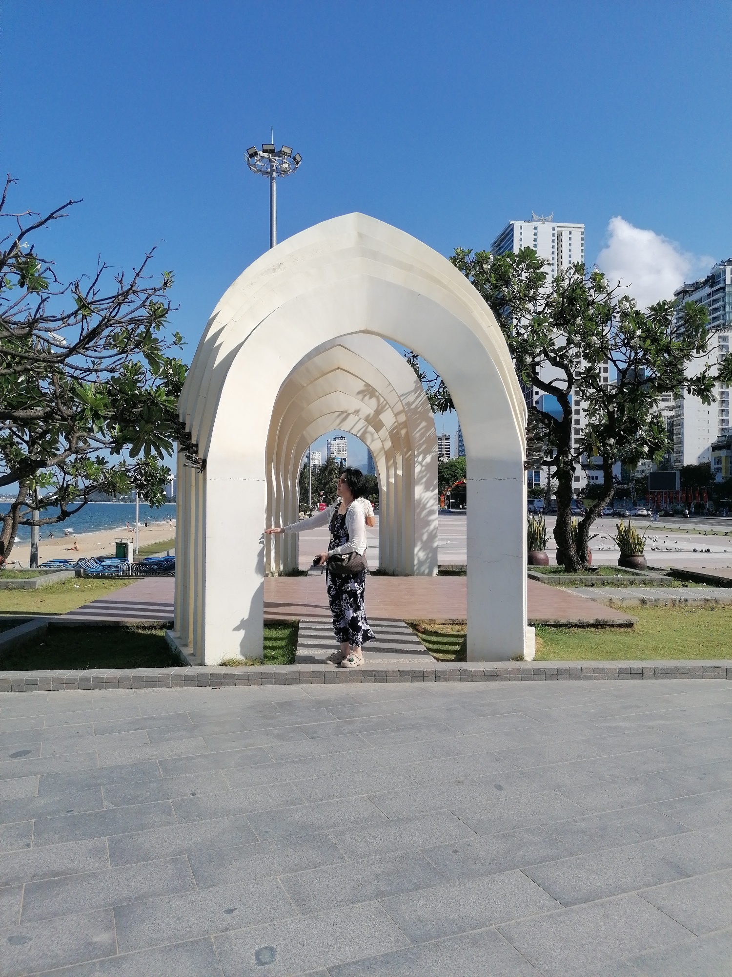 A woman stands within a series of white arches in Nha Trang Ward, Vietnam. She faces right, wearing a floral jumpsuit and white cardigan. Trees flank the arches, and a beach is visible to the left. In the background, buildings rise against a blue sky. A street with cars and pedestrians runs parallel to the arches. The foreground consists of a paved walkway.