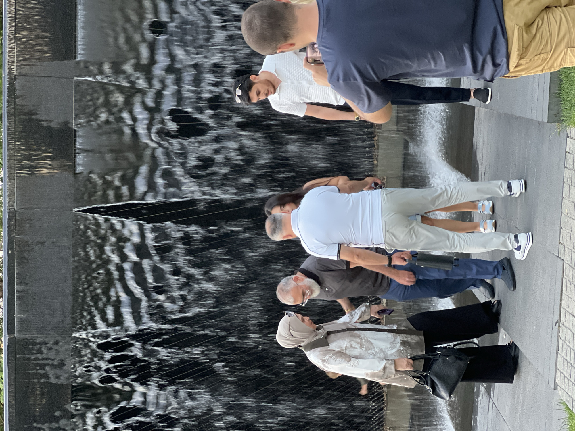 The image shows a group of people standing near a modern water feature, possibly a waterfall or a fountain. The water is cascading down a dark, textured surface, creating a dynamic visual effect. The setting appears to be an urban outdoor space, with a paved walkway or plaza surrounding the water feature.
Several individuals are visible, mostly in the foreground and midground, engaged in various activities. One person in the foreground is wearing a light-colored hijab and a patterned jacket, carrying a black handbag. To their right, another person in a dark grey shirt and blue pants is looking down, and further to the right, a person in a white shirt and light-colored pants is also looking towards the water. In the background, the backs of two individuals are visible, one in a dark blue shirt and tan shorts, and another in a white shirt. The presence of a mobile phone suggests people are taking photos or videos of the water feature.
The time of day is difficult to determine precisely, but the lighting suggests it is daytime. The weather appears to be pleasant, as people are dressed in casual, light clothing. There are no visible text elements in the image. The overall mood seems relaxed, with individuals observing and interacting with the water feature. The location cues are general to an urban park or public space with contemporary design elements.