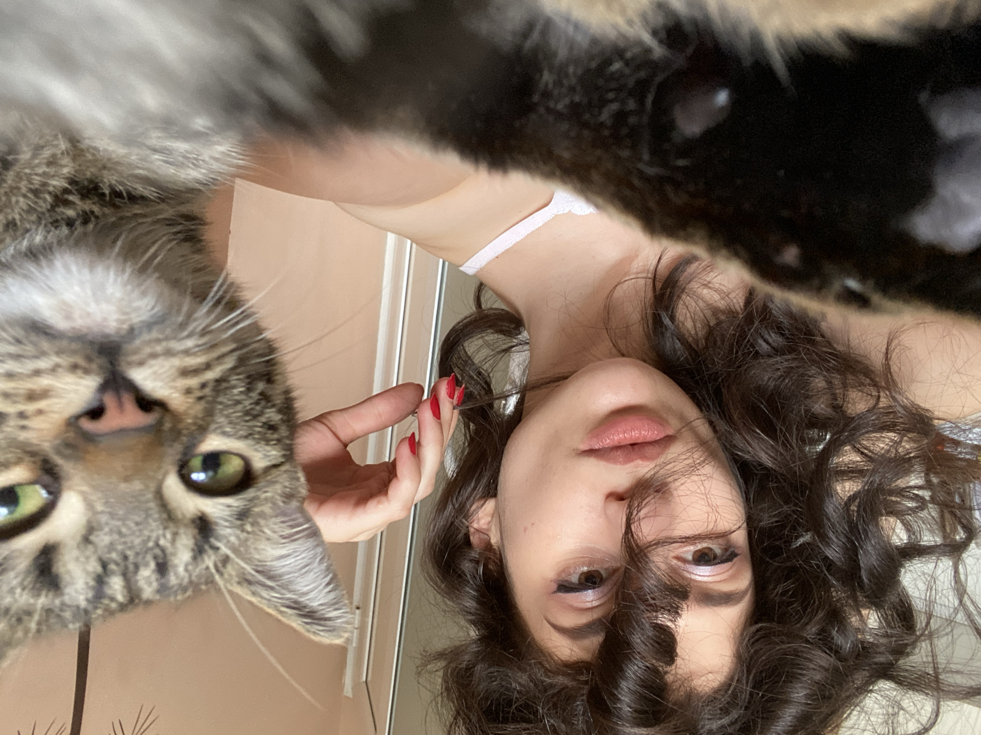 The image depicts a close-up selfie taken indoors, likely in a residential setting in Sofia, Bulgaria. The foreground is dominated by the blurry fur of a cat, obscuring much of the bottom portion of the frame. To the left and slightly behind the cat, a young woman is visible. She has dark, curly hair that frames her face, with some strands falling across her left eye. She is wearing light-colored clothing, possibly a camisole. Her right hand, adorned with red nail polish, is reaching towards her hair, holding a pair of small scissors, suggesting she might be trimming her hair. Her makeup includes eyeliner and eyeshadow. To the right, a tabby cat is partially visible, its face in sharp focus. The cat has large, green eyes and is looking directly at the camera. Its ears are perked, and whiskers are prominent. The background appears to be a wall with a subtle pattern and a doorway or partition on the right side. The lighting suggests an indoor environment with natural light coming from the left.