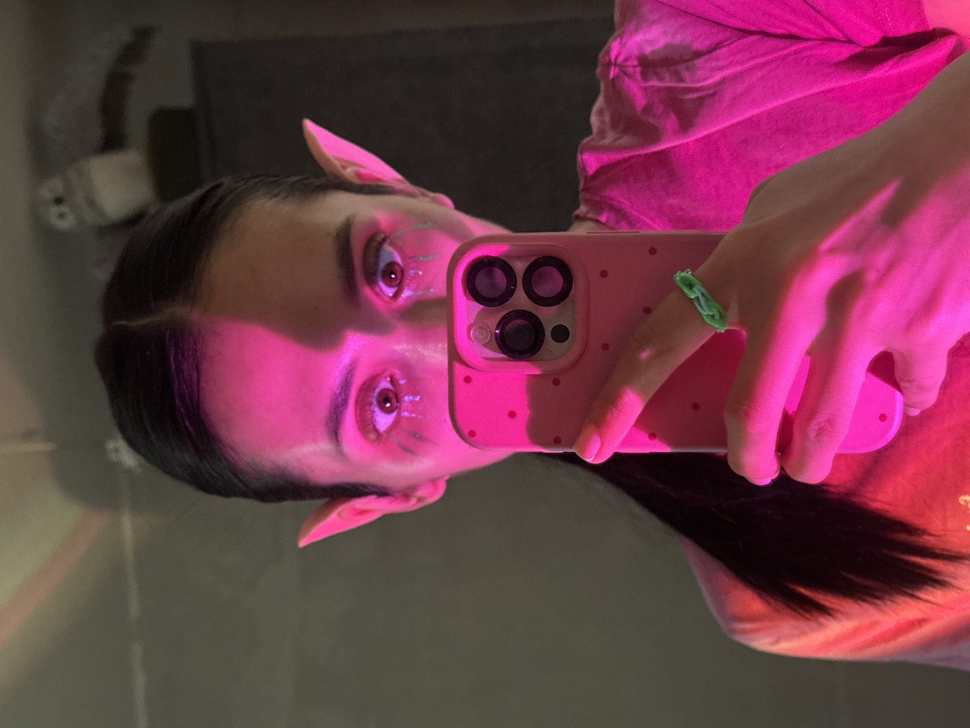 The image shows a person taking a selfie with a pink phone, illuminated by a bright pink light. The person has pointed ears, possibly as part of a costume or effect. There is makeup around their eyes. They are wearing a green rubber band ring. A blurry reflection, including a toilet paper dispenser, is visible in the background. There's no way to know if this image was taken in Votkinsk, Russia.