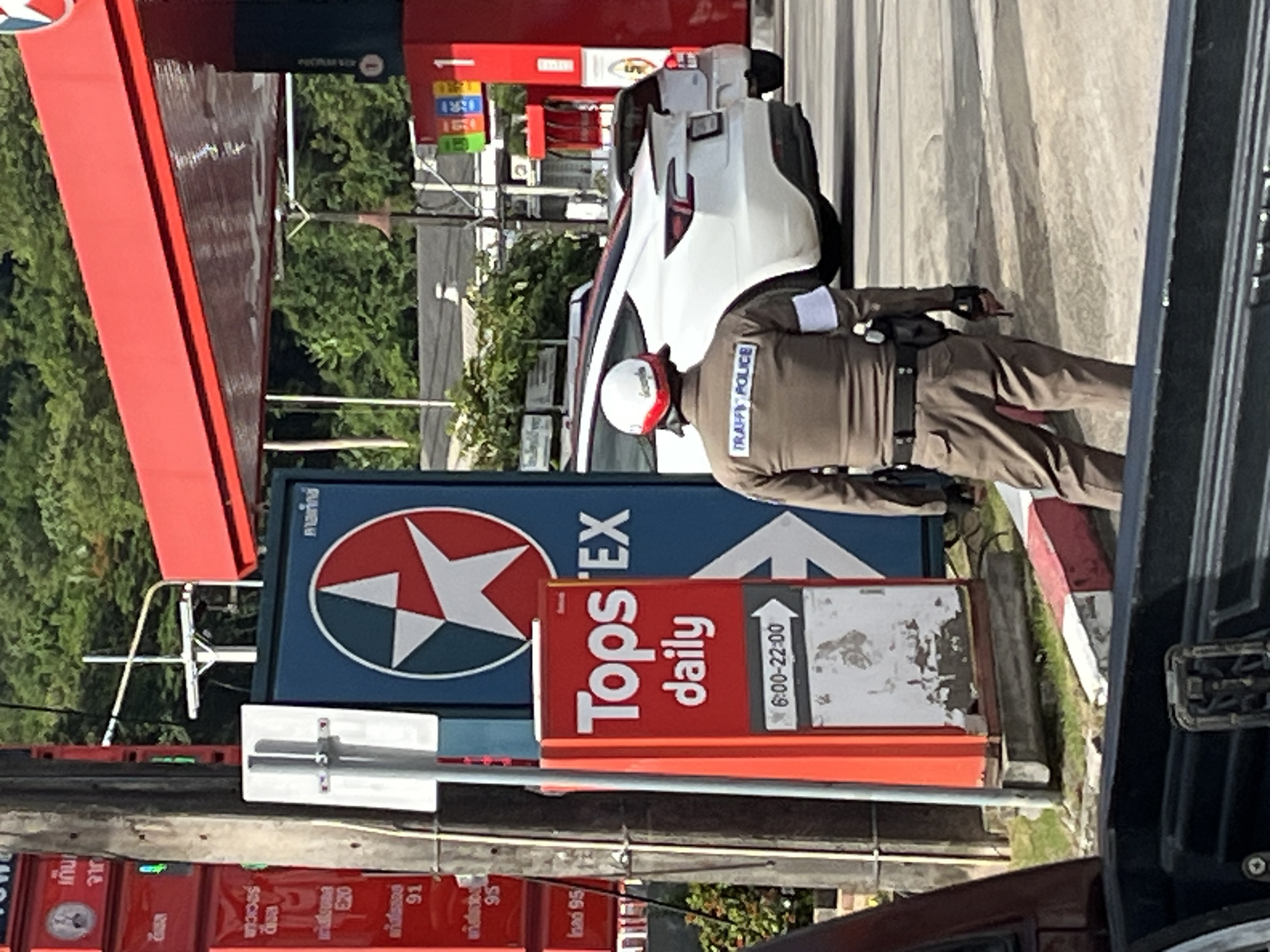 At a gas station in Phuket City Municipality, Thailand, a traffic police officer stands facing away from the camera, observing a white Tesla. The officer is wearing a brown uniform with a white armband and a helmet. The gas station features red and blue signage, including a large Exxtra logo. A red "Tops Daily" sign is also visible. The environment appears to be outdoors with trees in the background, suggesting a sunny day.