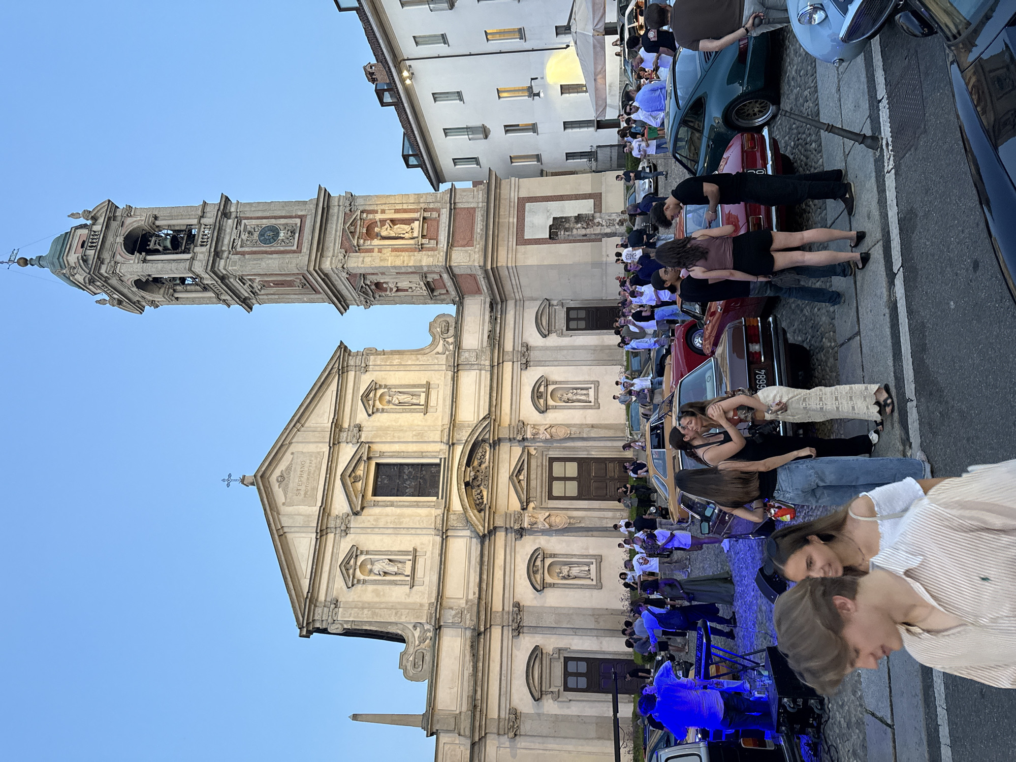 The image shows a street gathering in front of a church and a bell tower in Milan, Italy. A crowd of people, dressed in casual summer attire, are mingling and observing a collection of vintage cars parked on a cobblestone street. The church, with its neoclassical facade and statues, dominates the background, while the tall, ornate bell tower stands to its right. The sky is clear and blue, indicating a late afternoon or early evening setting. Some individuals are interacting with each other, while others are looking at the cars or listening to music, possibly from a DJ setup visible on the left. The overall atmosphere appears festive and relaxed.