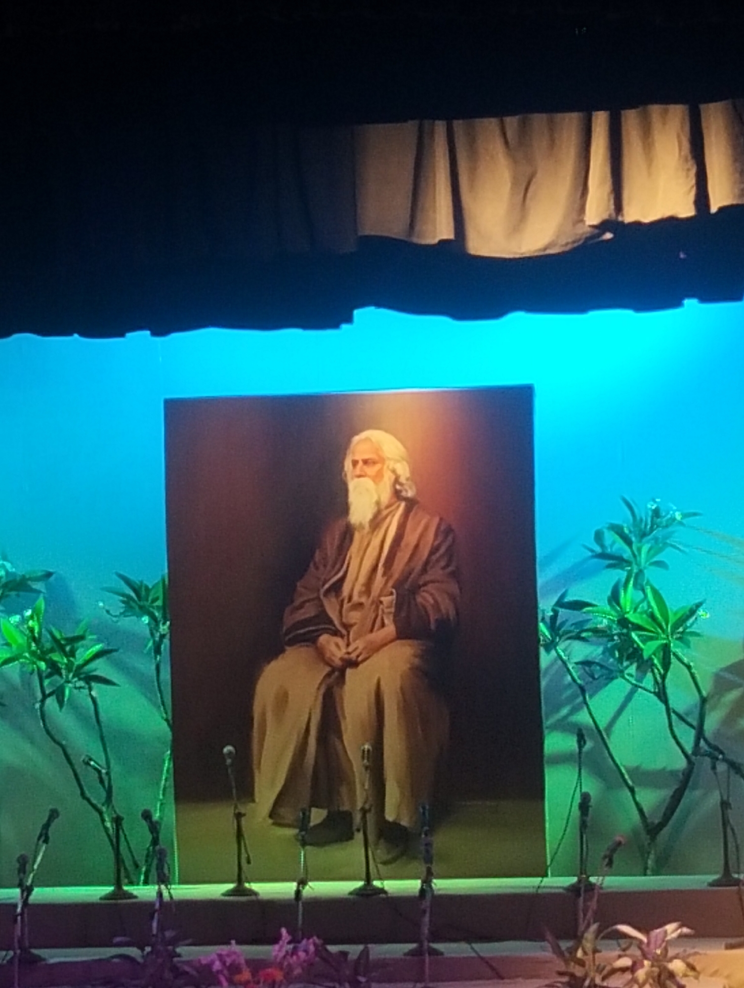 A full shot shows a stage with a large portrait of a man with white hair and a beard, seated and wearing a robe. The stage is decorated with greenery, and several microphones are arranged in front of the portrait. A curtain hangs above the stage. The location is Unknown, Unknown.