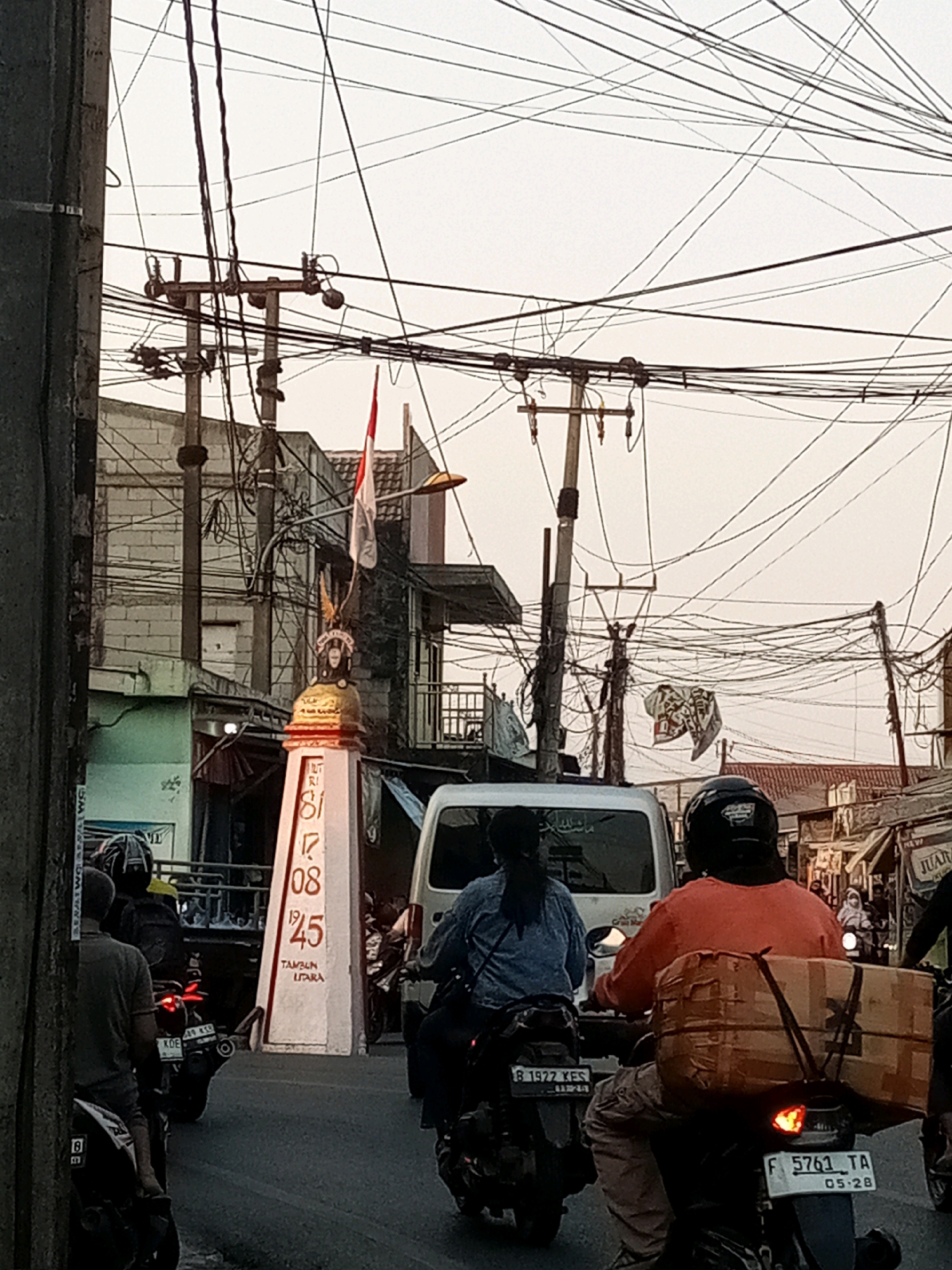 This is a medium shot, captured at an eye-level perspective, of a street scene in Karangsatria, Indonesia. The sky is overcast and pale. In the foreground, a concrete pole is visible on the left. A monument-like structure stands in the middle, bearing the dates "18170825" and "TAMBUN UTARA" in red numerals and text, respectively. A flag, red and white, is affixed to the top of this monument. Several motorcycles are on the road. One motorcycle, with a license plate reading "F 5761 TA," is carrying a large cardboard box secured to its rear. Another motorcycle, with a license plate reading "B 1922 KES," is in front of it. Behind the monument, a white van is visible. The background is filled with numerous electrical wires strung between poles, typical of a densely populated urban area. Buildings line the street, some appearing to be shops or residences. The overall impression is one of a busy street on a cloudy day.