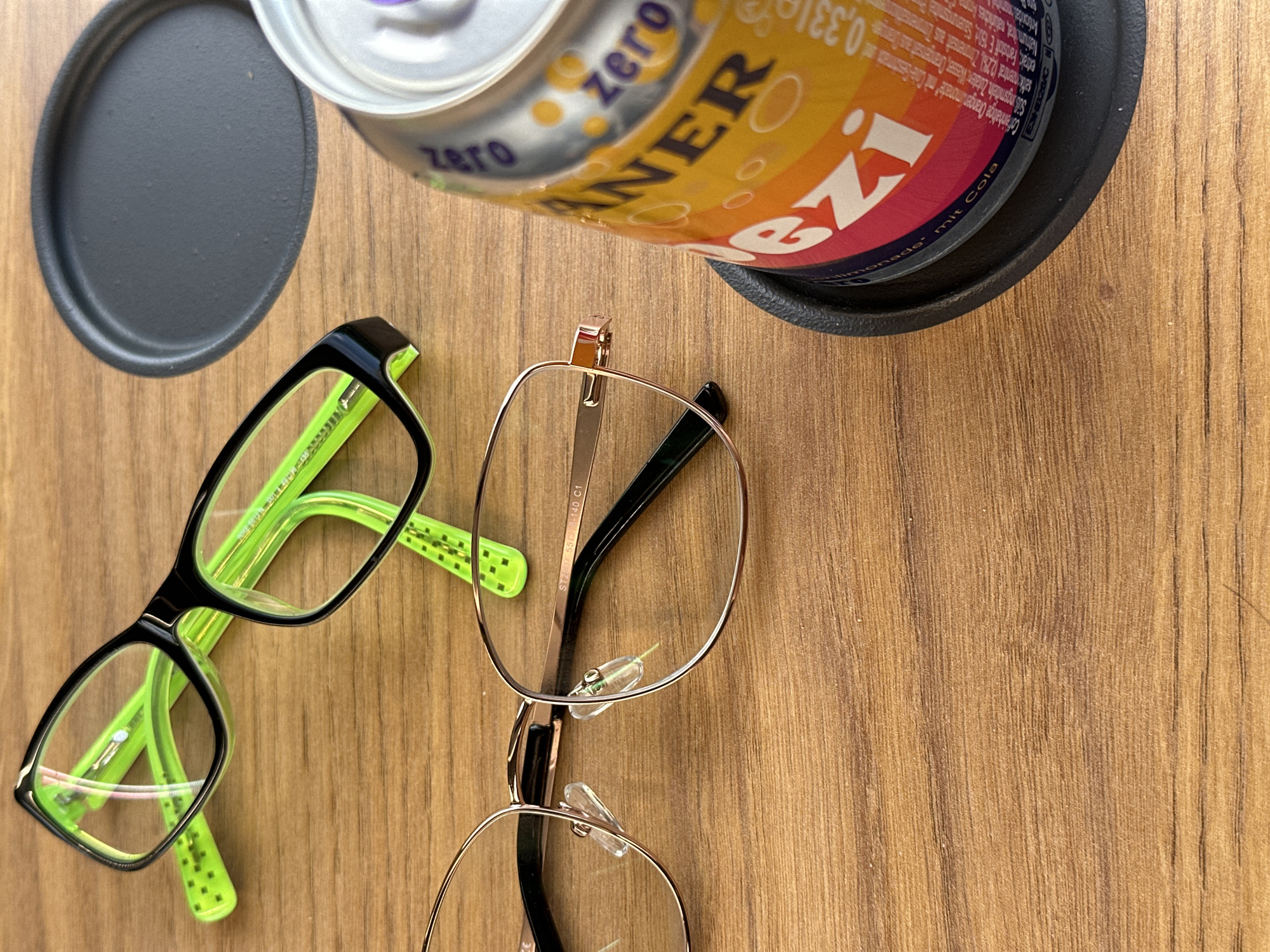 This is a close-up, overhead shot of two pairs of eyeglasses and a can of "ALLSEITS Zero" orange and lemon soda with cola flavor, all resting on a wooden surface. One pair of glasses is black with neon green arms, featuring a lime green pattern on the arms. The other pair is rose gold with clear nose pads. The soda can is partially visible, with its label showing bold, colorful text including "ZERO", "ANER", and "0,33l". The text on the can is in German and describes the ingredients and flavor profile of the beverage. A dark gray coaster is also visible beneath the can. The lighting suggests it could be daytime, possibly outdoors, given the natural wood grain and the casual arrangement of objects. There are no people or animals present in the image. The overall mood is casual and relaxed, hinting at a moment of pause or refreshment.