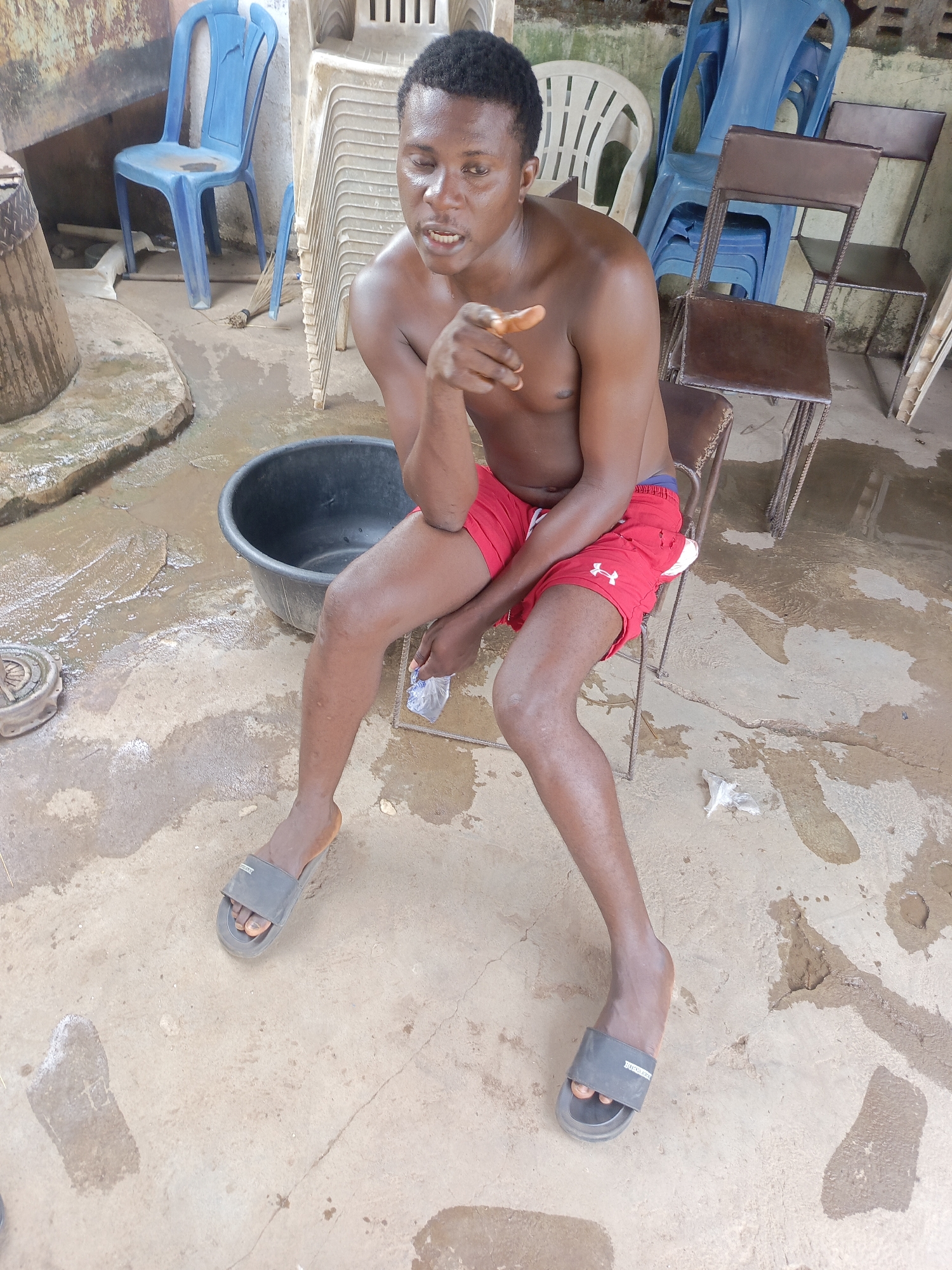 A shirtless man wearing red shorts and gray slide sandals sits on a chair outdoors. The man is pointing toward the viewer. A black plastic basin sits near him. The ground is concrete and appears wet. Stacks of plastic chairs and metal chairs are visible in the background. The location is indicated as Unguwar Zaria, Nigeria. A piece of clear plastic is on the floor near the man's leg.