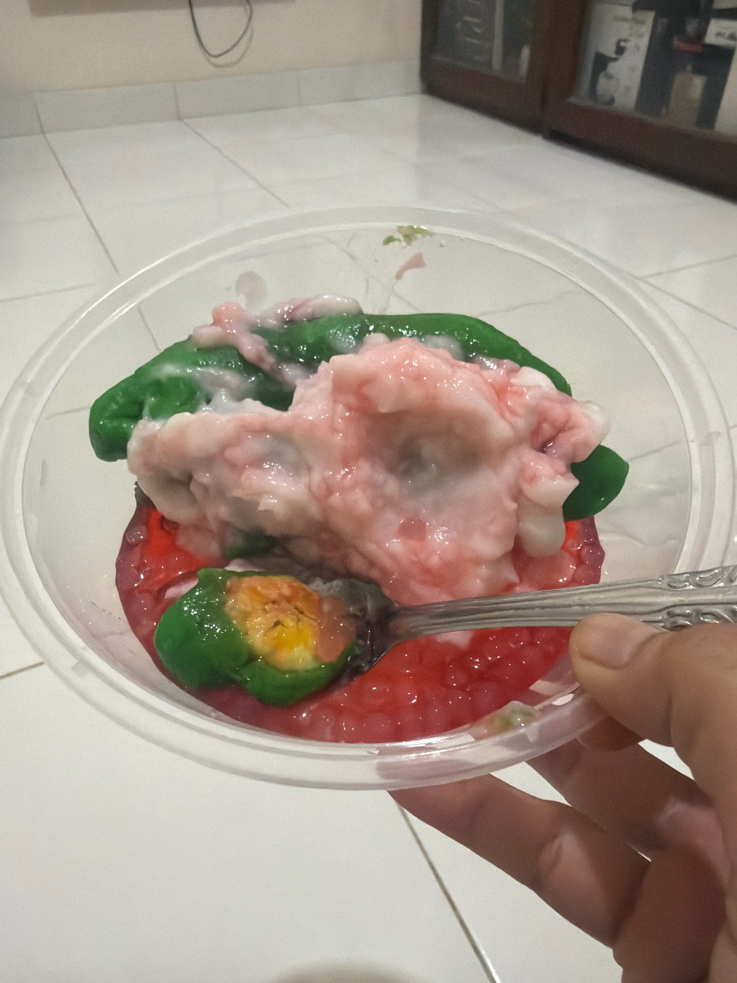 The image shows a close-up, overhead view of a clear plastic container filled with a colorful dessert. The dessert consists of green chewy pieces, a pinkish-white creamy sauce, and a red jelly-like substance at the bottom. A spoon is visible, scooping up a portion of the dessert, revealing more of the green and yellow interior of one of the chewy pieces. The container is placed on a tiled floor, which appears to be white with gray grout lines. In the background, a wooden cabinet with glass doors is visible, displaying various items. The lighting suggests an indoor environment, possibly a home or a restaurant. The image does not provide any specific information about the city of Balikpapan, Indonesia, or any context that would place the scene there.