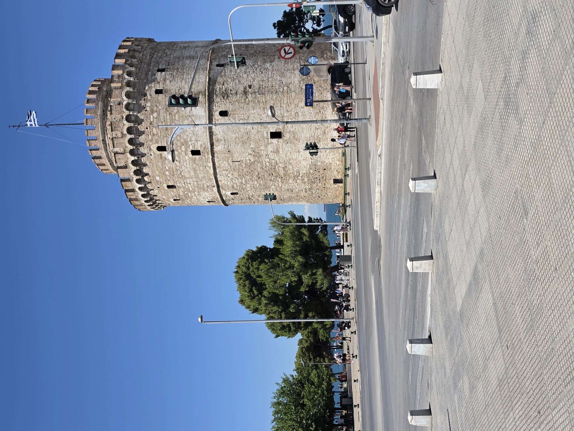 The image shows the White Tower, a historical monument in Thessaloniki, Greece. The tower is a prominent cylindrical stone structure with crenellated battlements and small windows. A Greek flag flies from a flagpole atop the tower. In the foreground, a paved plaza with concrete bollards is visible, bordering a street intersection. Traffic lights, streetlights, and road signs are present. People are seen walking and standing near the tower and along the waterfront in the background, where trees line the promenade. Several vehicles, including cars and motorcycles, are on the street. The sky is clear and blue, indicating a sunny day.