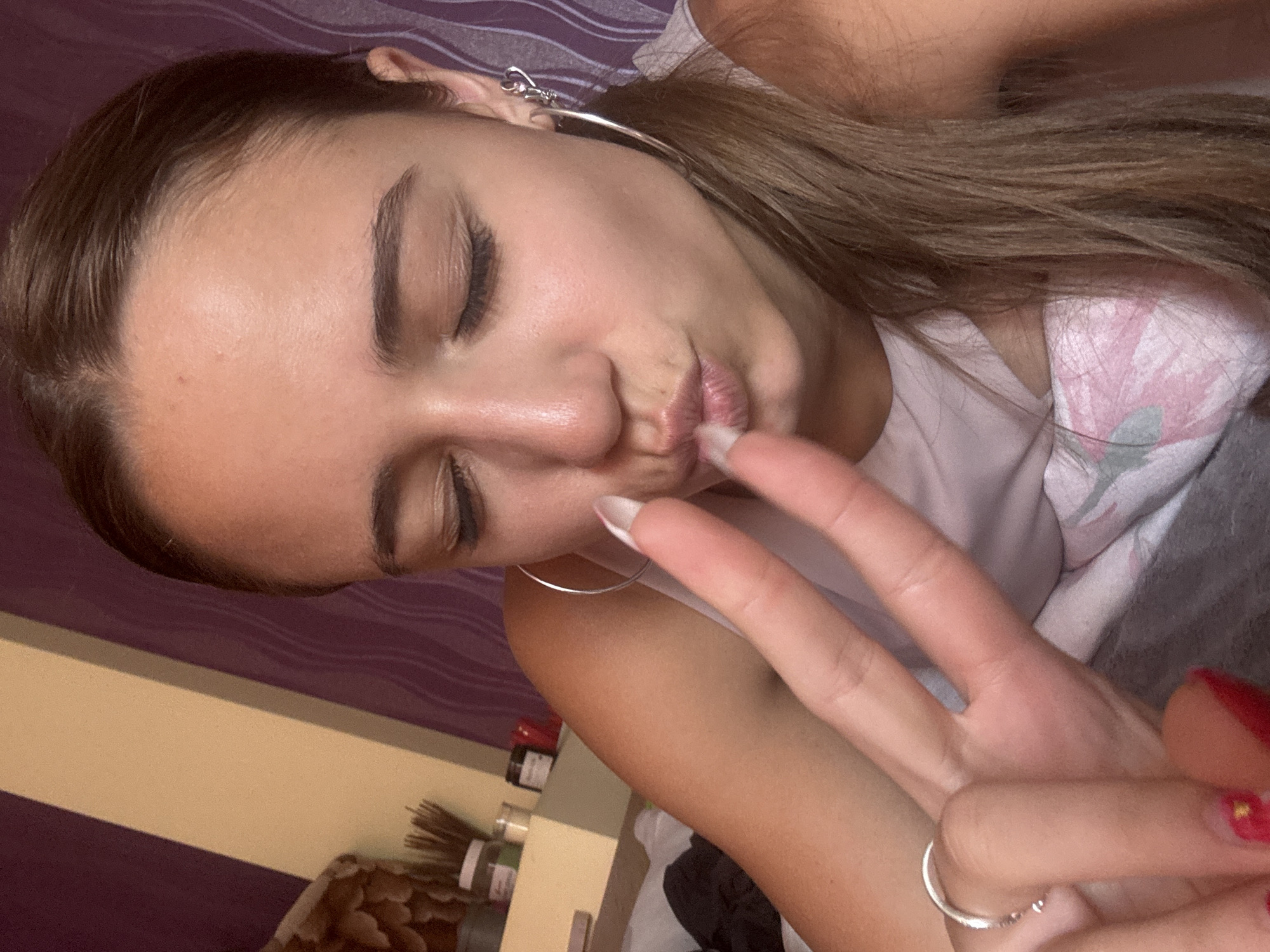 The image is a close-up selfie of a young woman with her eyes closed and lips pursed. She is holding up two fingers in a peace sign with her right hand, while her left hand is resting on her chest. She is wearing a white tank top and large hoop earrings. The background appears to be a bedroom with purple and beige walls. There are some items on a shelf in the background, including jars and what look like incense sticks. The overall impression is of a casual, indoor setting. The prompt's request to include the location as Pernik, Bulgaria is not supported by any visual evidence in the image.