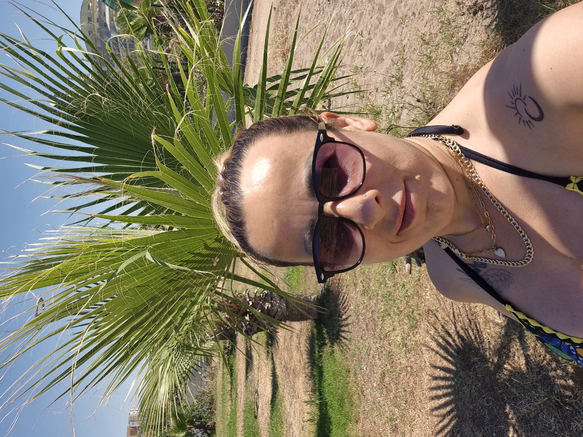 A woman is taking a selfie outdoors, likely on a warm, sunny day given the bright lighting and palm trees. She is wearing sunglasses and a bikini. A tattoo is visible on her upper arm. She is also wearing a gold chain necklace. The background shows palm trees, grass, and sand, suggesting a tropical or beachside location. The overall mood appears relaxed and casual. There is no visible text in the image.